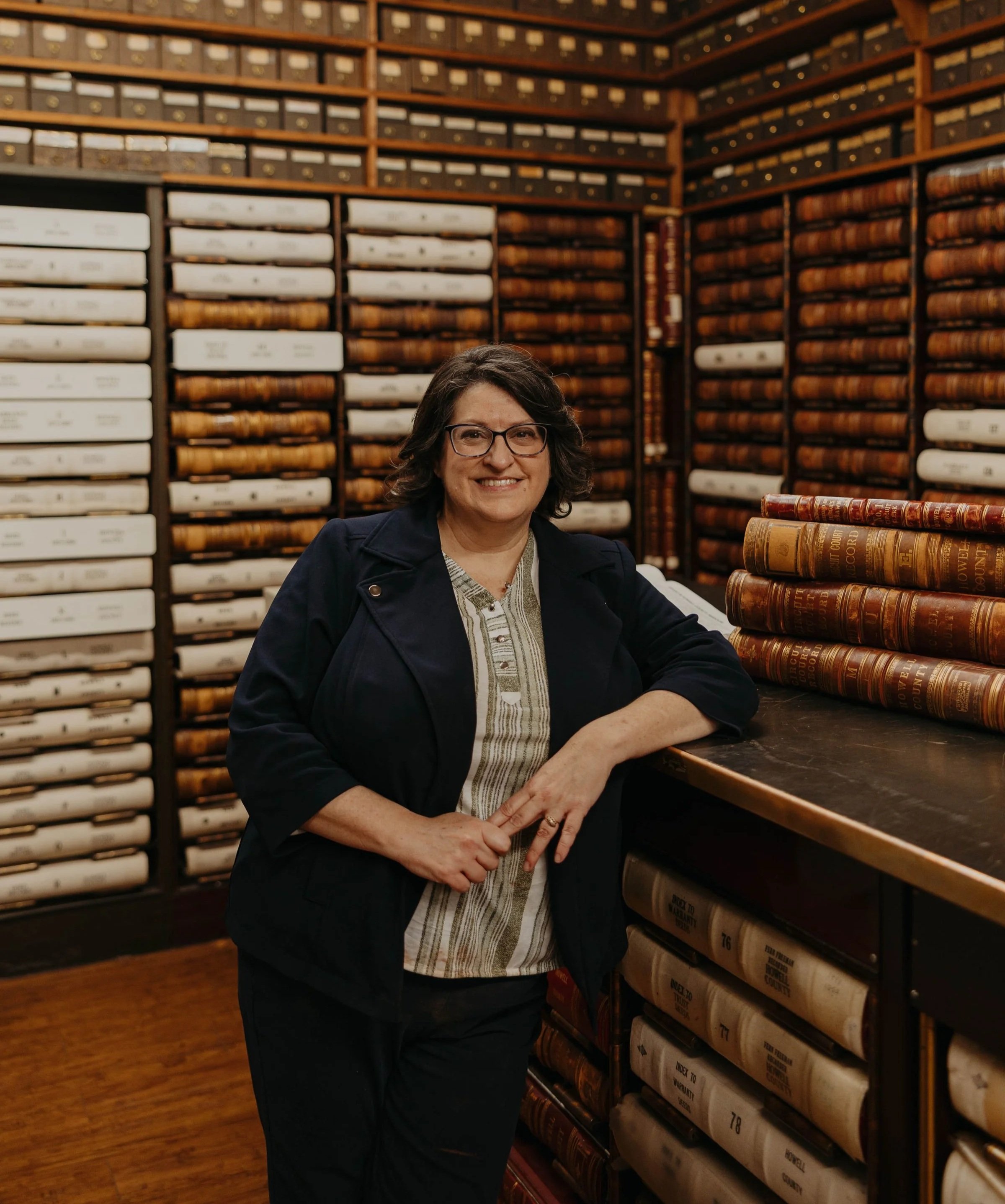 A woman with glasses and short dark hair smiling, standing in an archive room filled with shelves of legal or historical books.