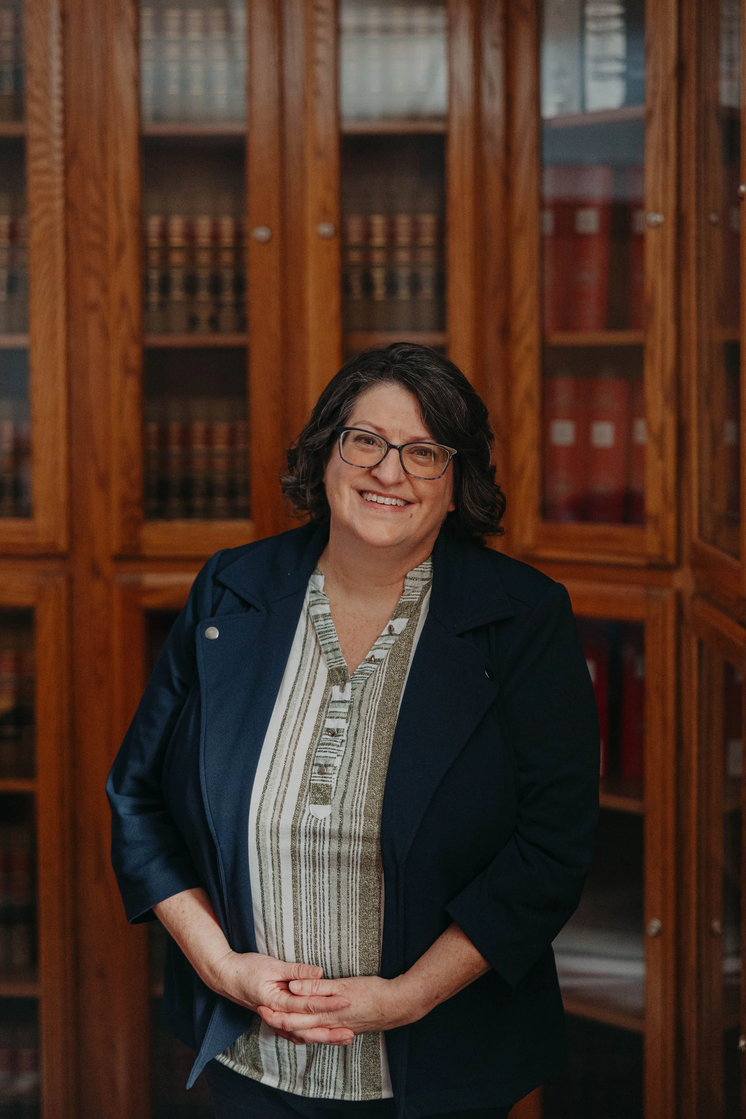 A middle-aged woman with short dark hair and glasses smiling, standing in front of a wooden bookshelf filled with law books.