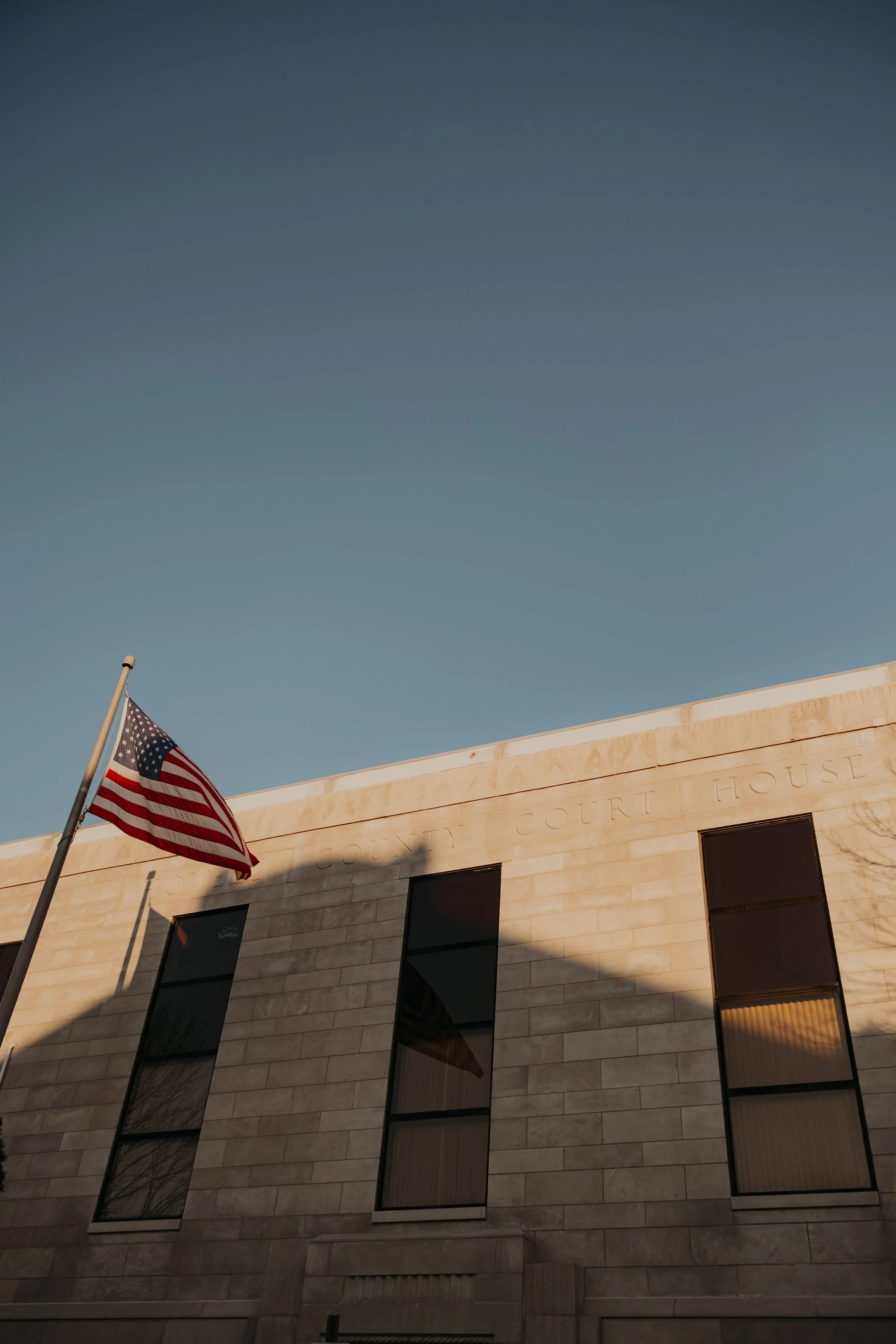 The image shows a beige courthouse building with dark-tinted rectangular windows. An American flag is flying on a flagpole in front of the building, waving in the wind. The sky above is clear with a gradient from darker to lighter blue.