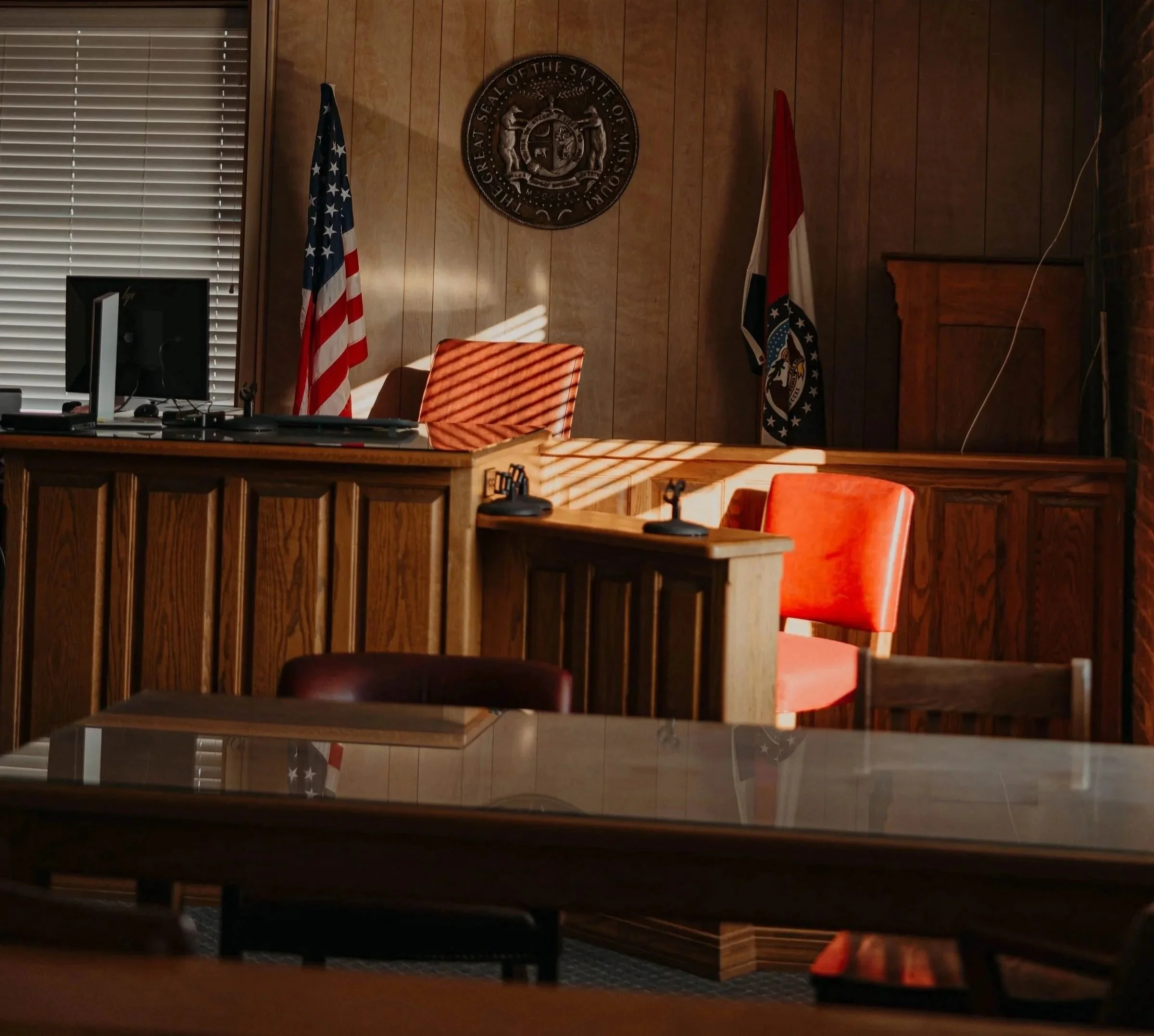 A courtroom or official government room with a wooden backdrop, two flags (United States and Missouri), a seal of the State of Missouri, a computer monitor, a wooden desk with papers, orange chairs, and sunlight casting shadows through blinds.