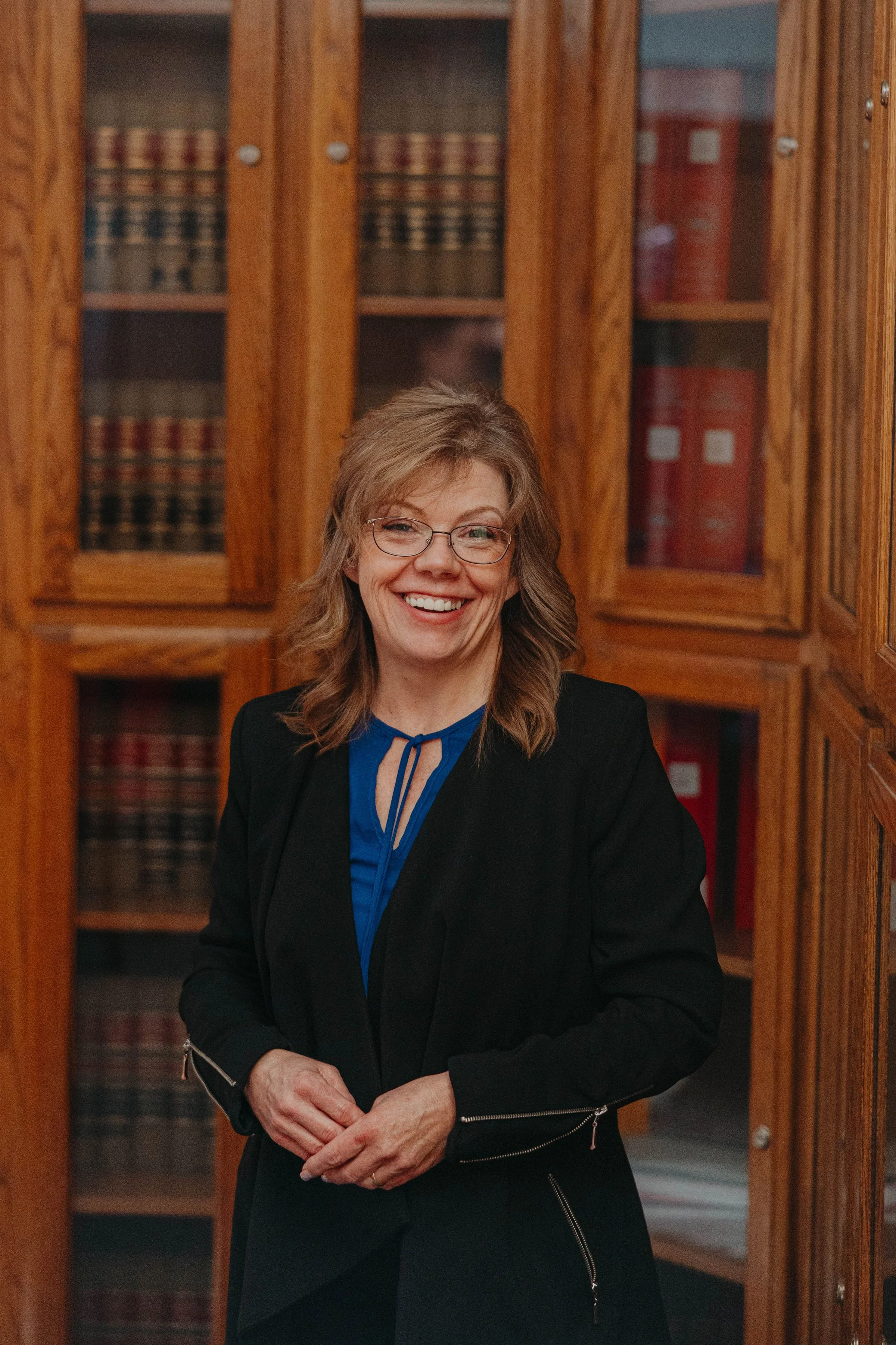 A smiling woman with glasses and wavy shoulder-length hair, wearing a black jacket over a blue blouse, standing in front of wooden bookshelves filled with legal books.