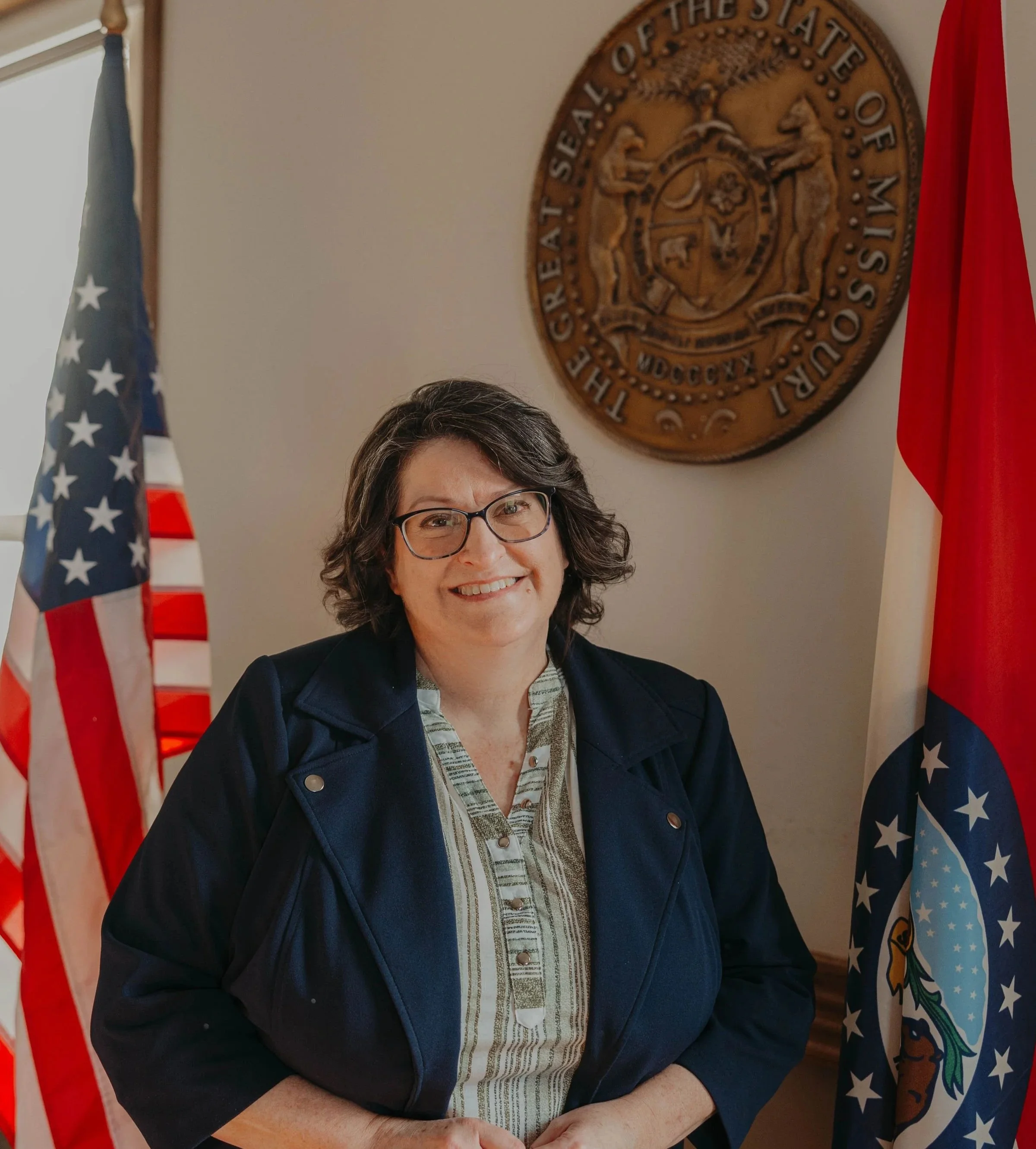 A woman with glasses smiling in front of an American flag, a state seal on the wall, and a red flag.
