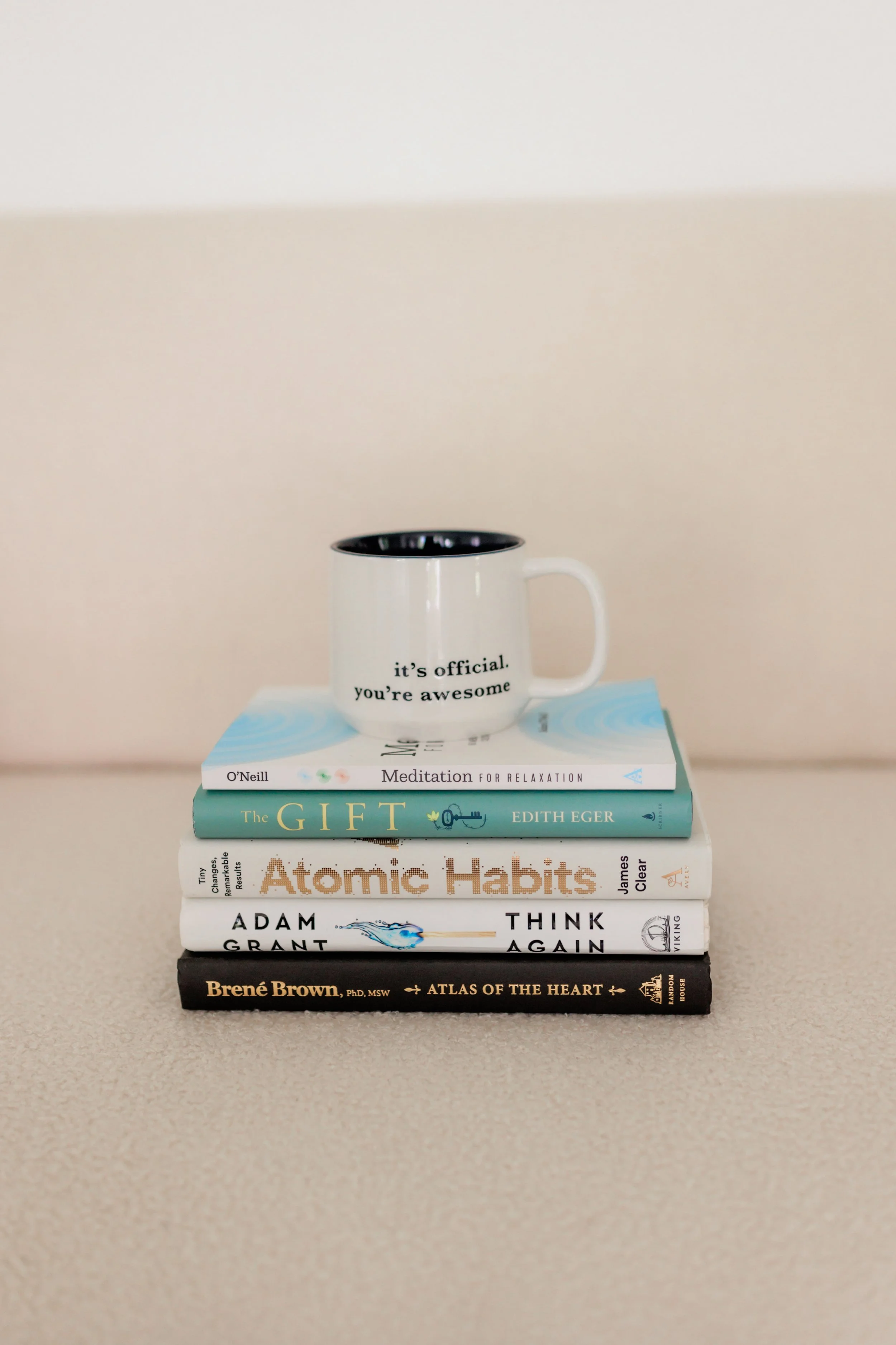 Stack of five books with a white mug on top, on a beige surface. The mug has the text 'it's official. you're awesome'.