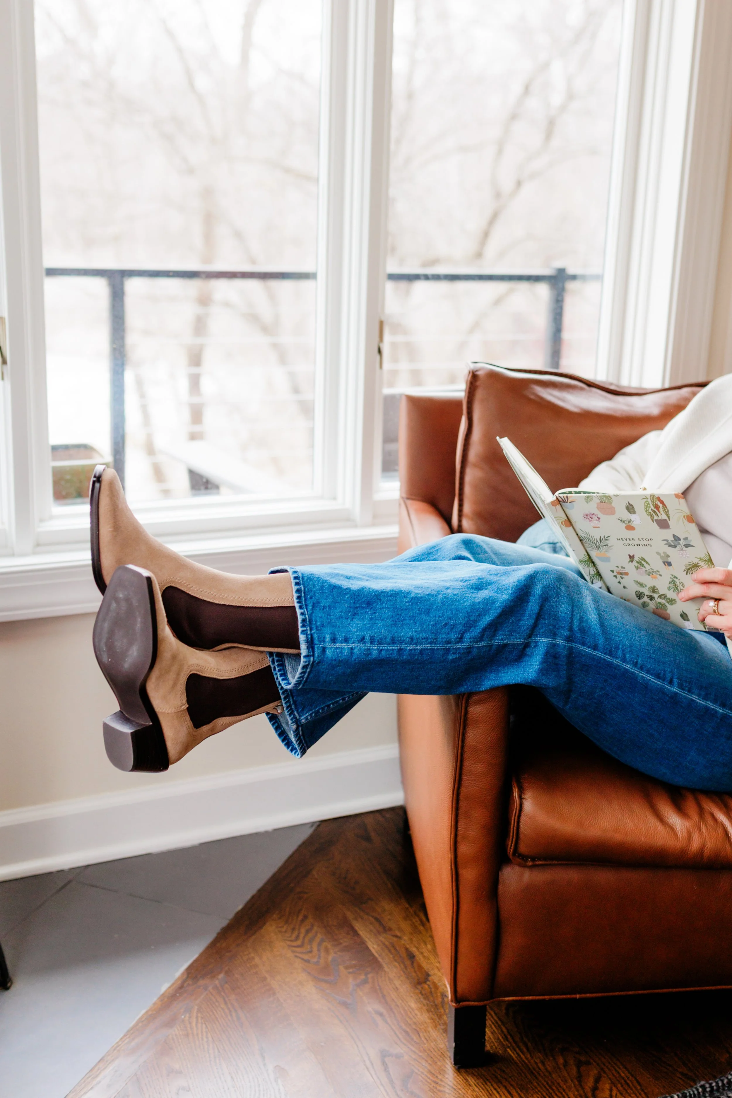 Person sitting on a leather couch with legs extended, reading a book, wearing jeans and beige cowboy boots, near a large window with a balcony outside.