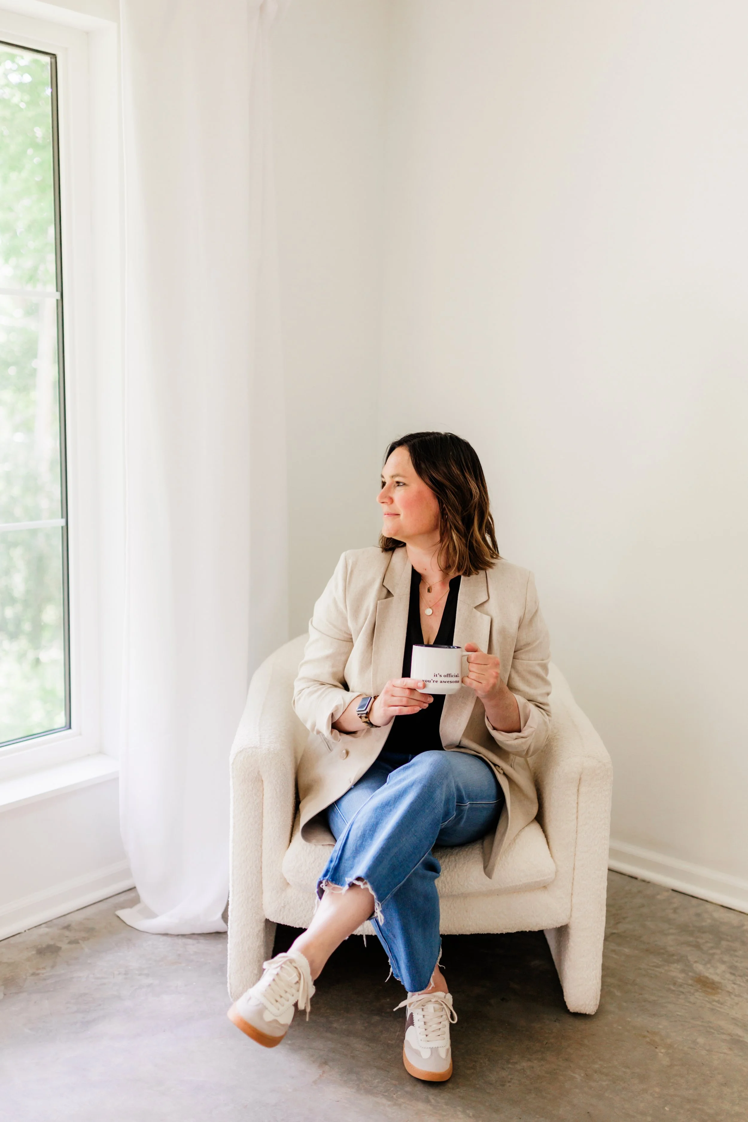 Woman sitting on a white armchair near a window, holding a mug, wearing a beige blazer, black shirt, blue jeans, and sneakers, looking out the window.
