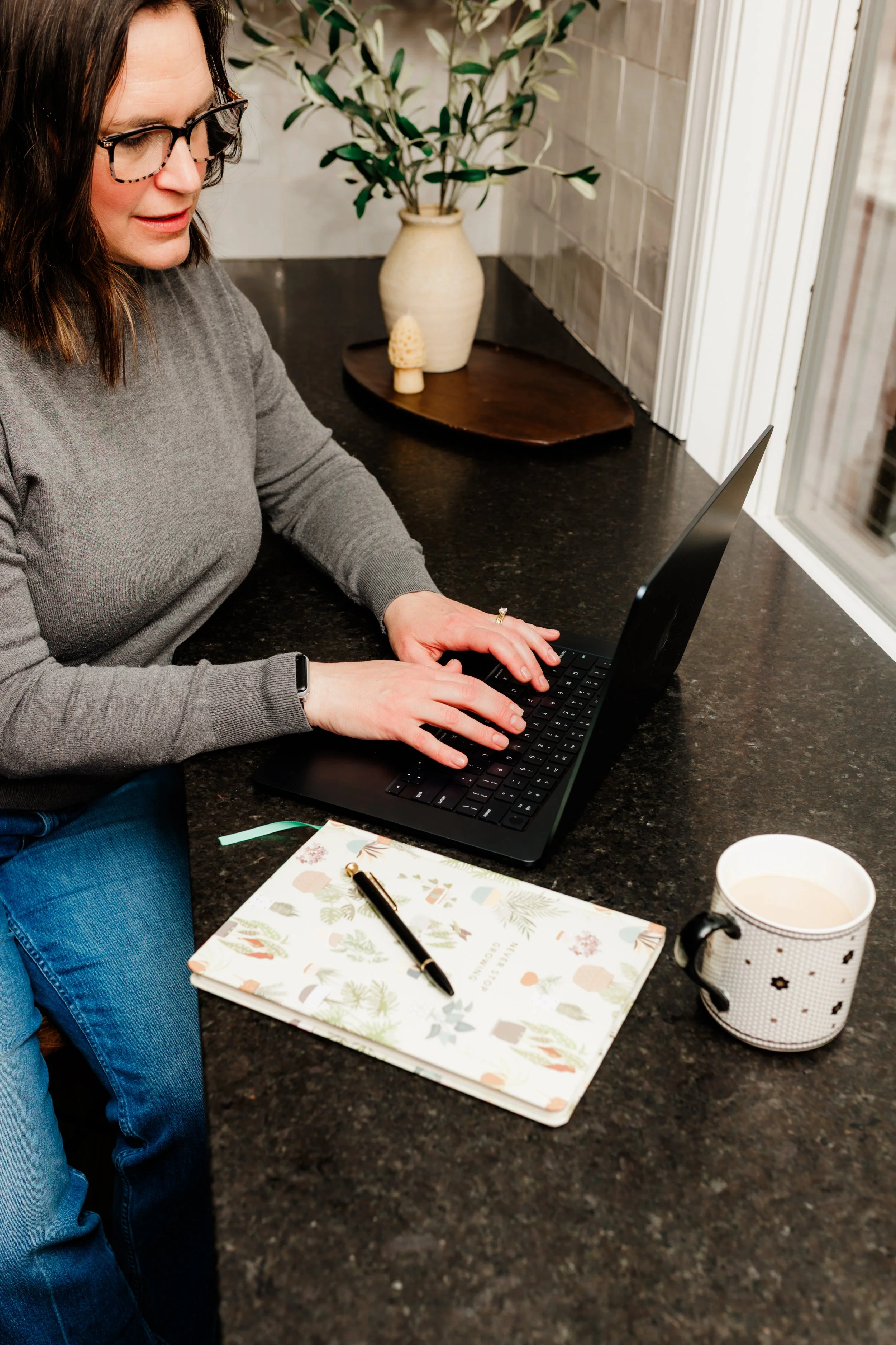 A woman in glasses using a black laptop at a kitchen counter with a notebook, pen, and mug nearby.