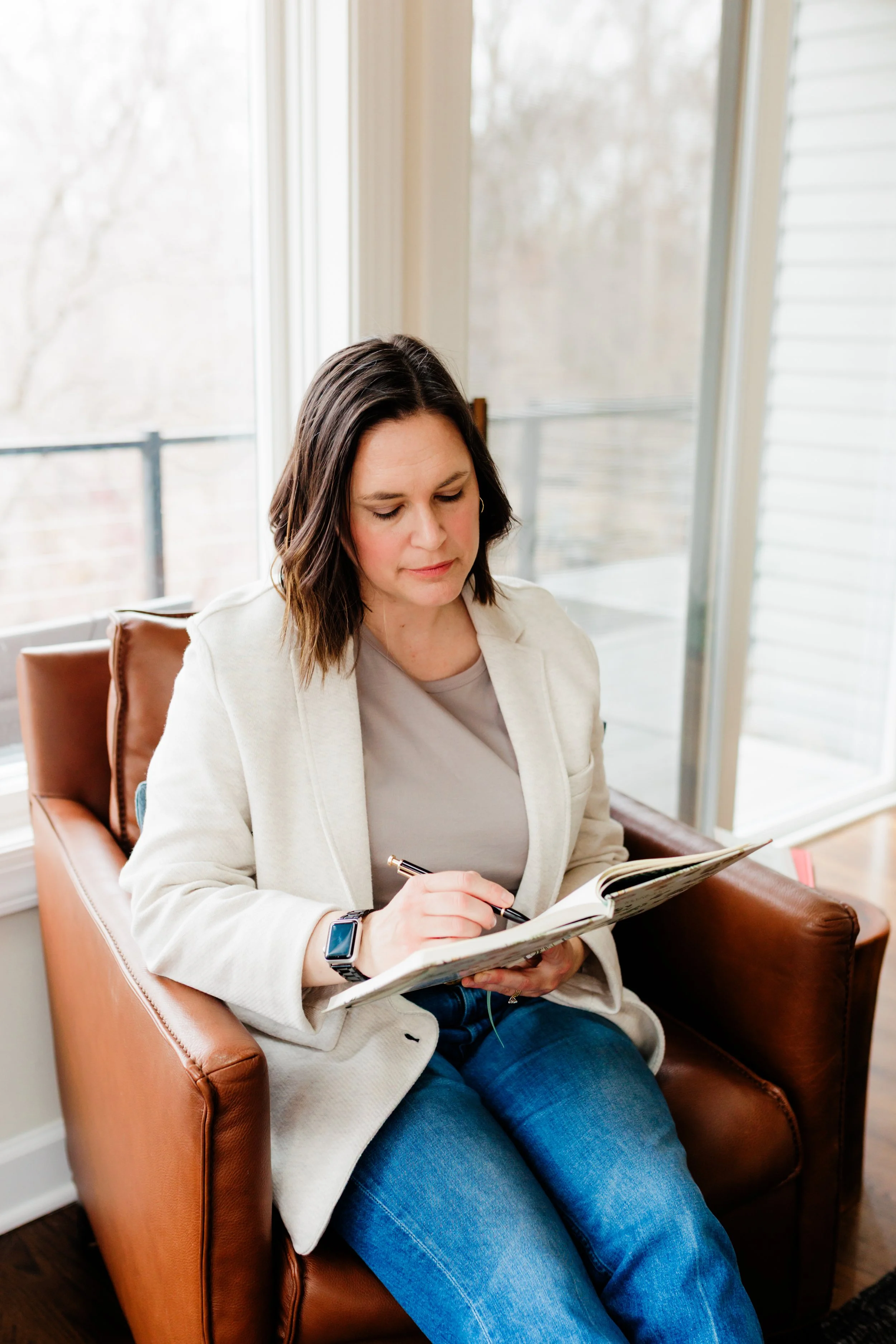 A woman with shoulder-length dark hair, wearing a beige blazer and jeans, sitting on a brown leather armchair in front of a large window, writing in a notebook with a pen.
