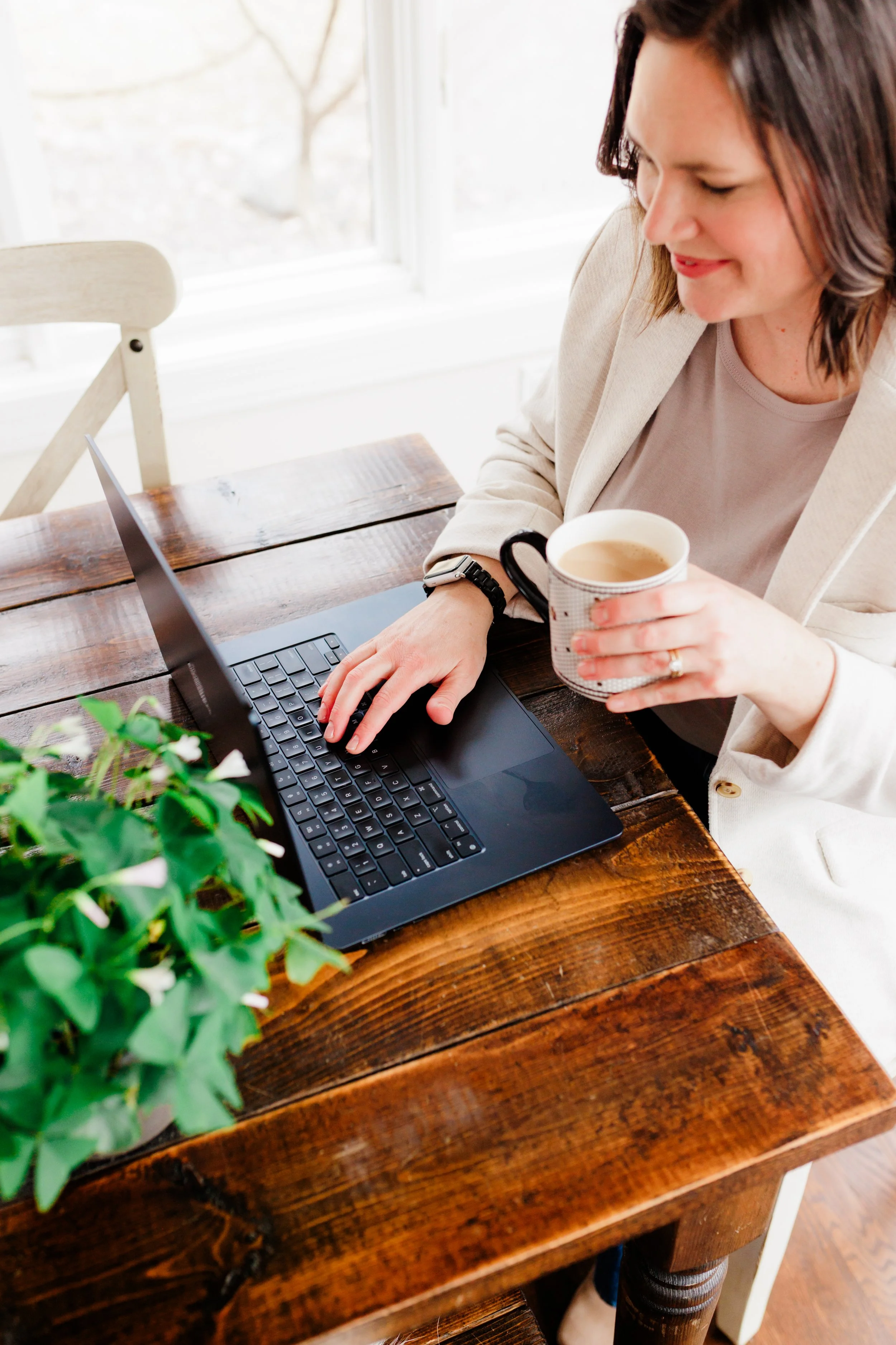 A woman sitting at a wooden table, working on a laptop, holding a mug, with a window and greenery in the background.