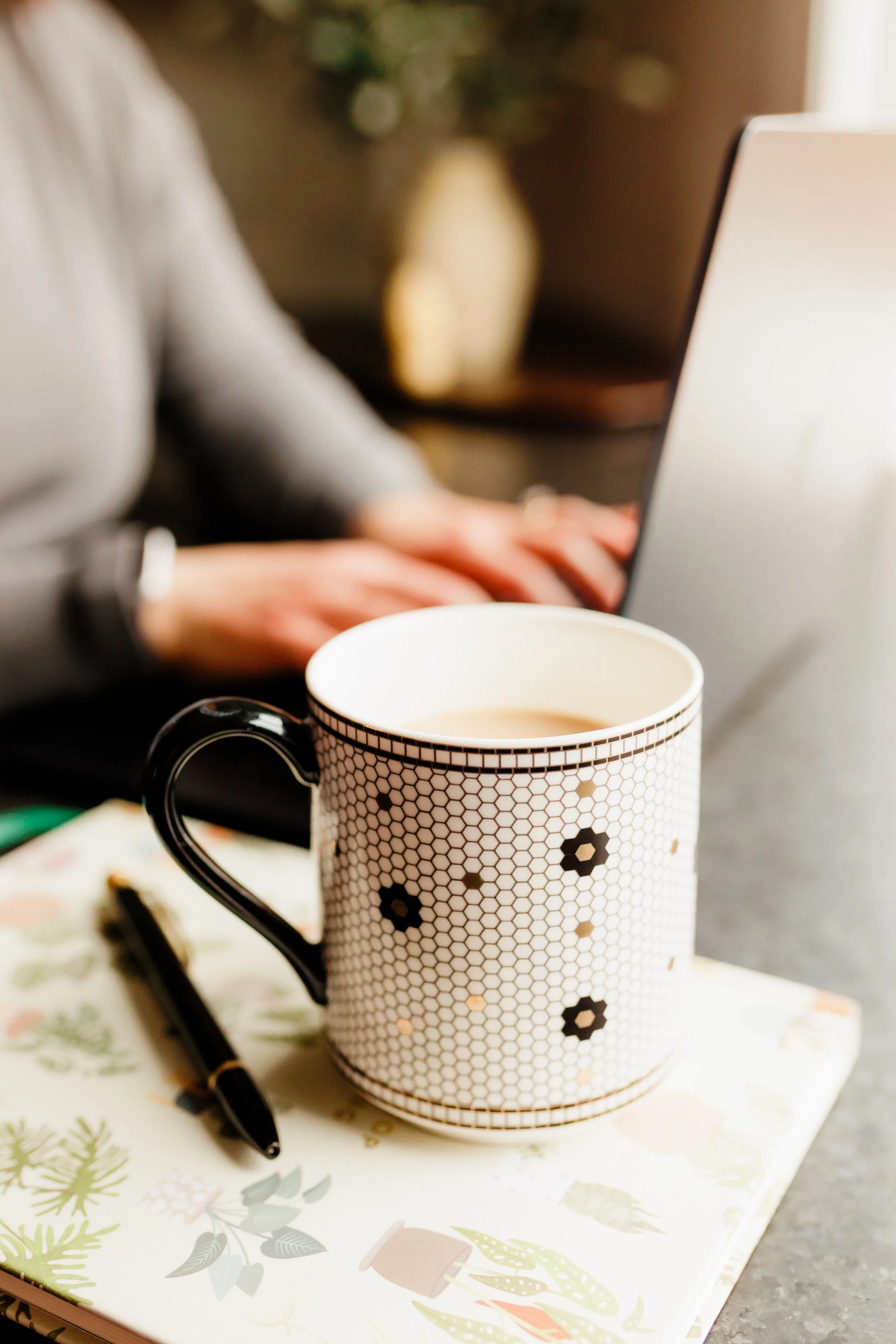 A patterned mug filled with coffee on a desk, with a notebook and pen beside it, and a blurred person using a laptop in the background.