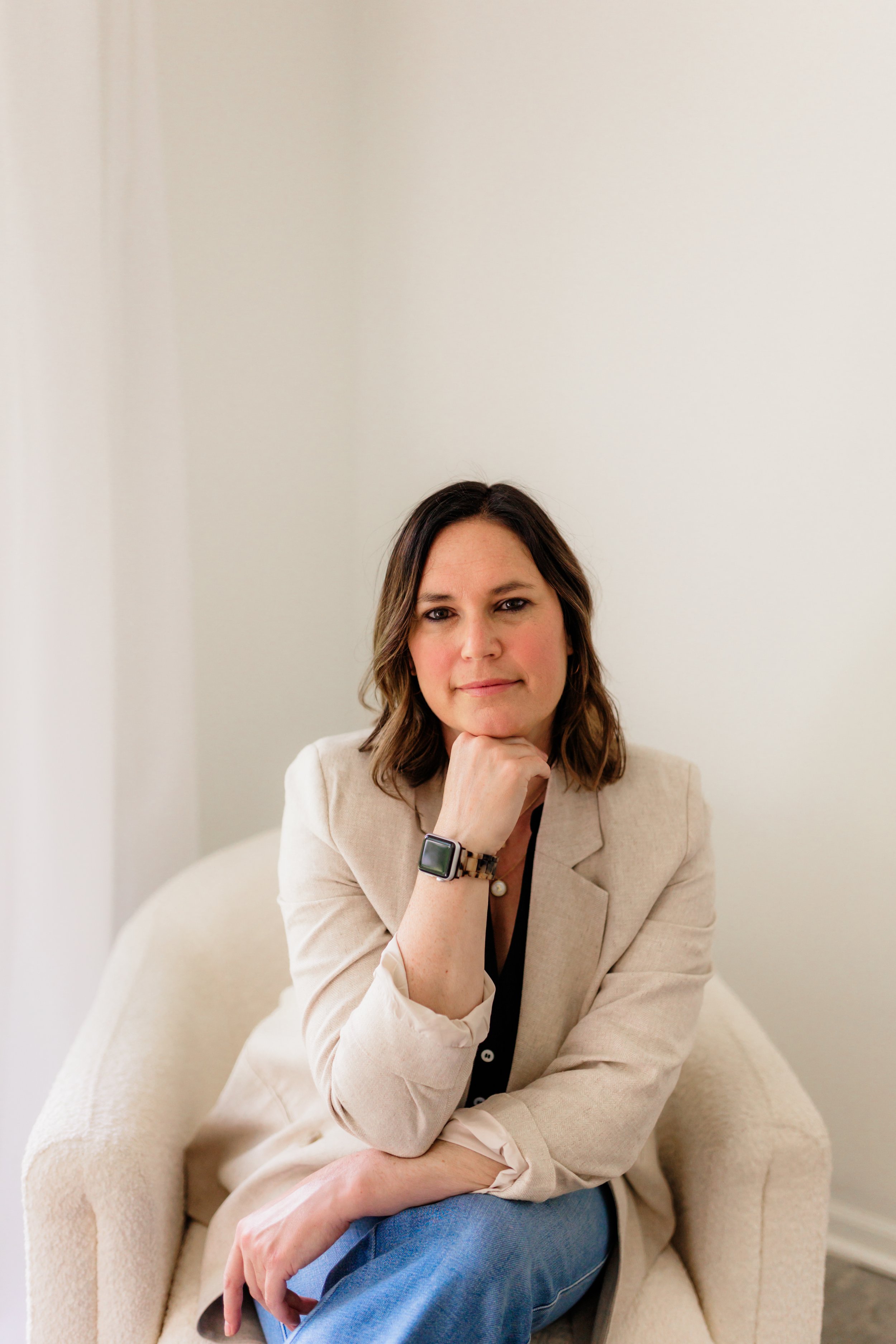 A woman with shoulder-length brown hair sitting on a cream-colored armchair, wearing a beige blazer, black top, blue jeans, and an Apple Watch, resting her chin on her hand and looking at the camera.