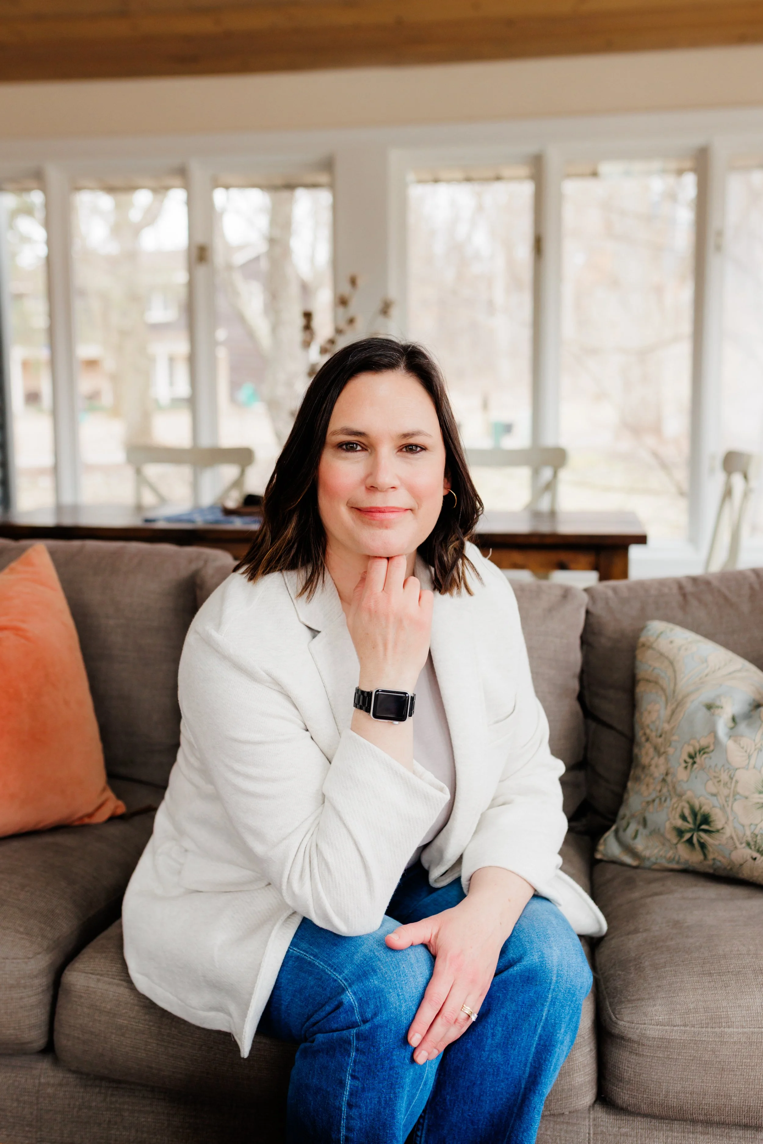 A woman with shoulder-length dark hair wearing a white blazer and blue jeans sitting on a beige couch in a bright room with large windows and a wooden table in the background.