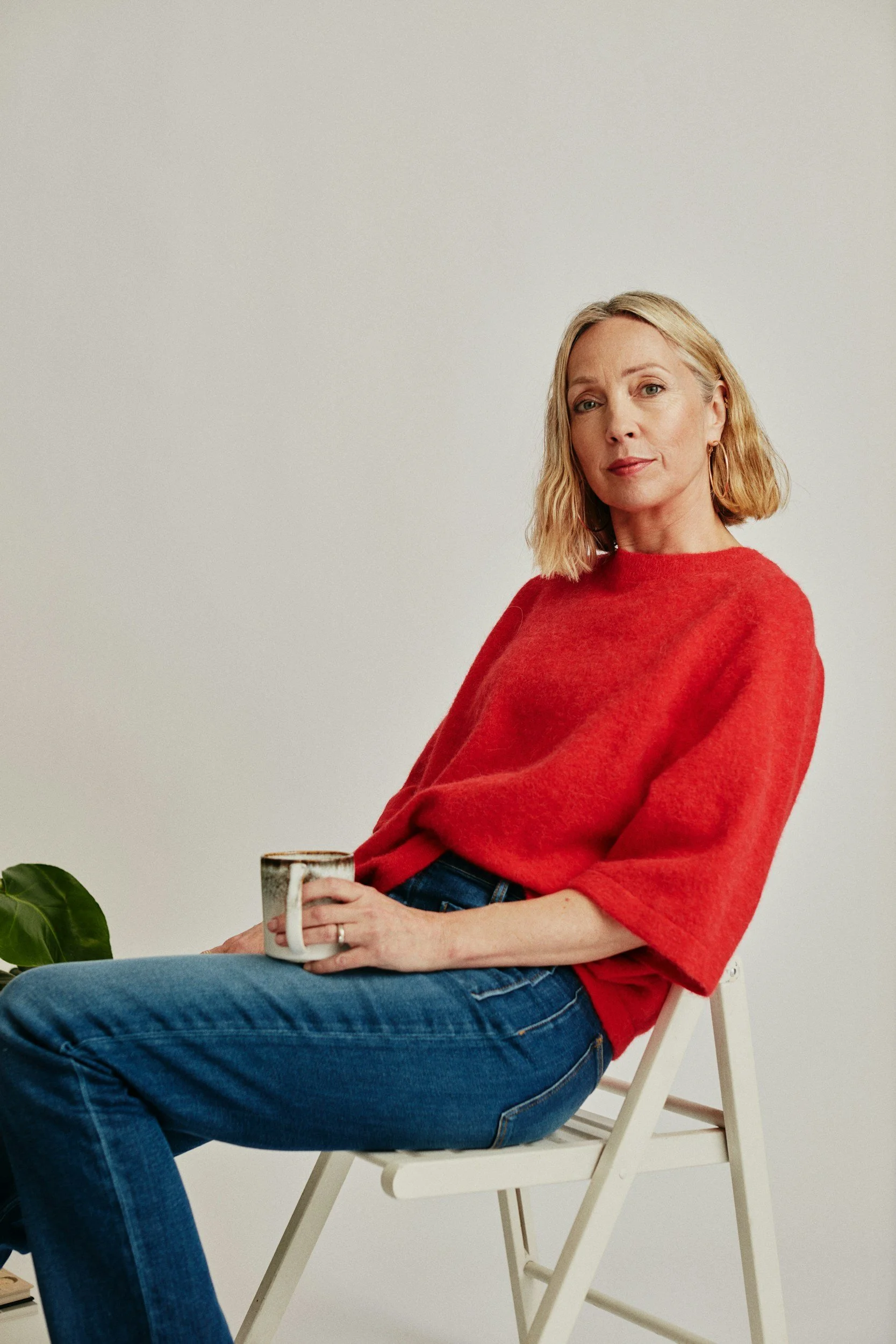 A woman with blonde hair wearing a red sweater and blue jeans sitting on a white chair, holding a mug with a colorful design, in front of a plain light-colored wall with a plant on the lower left.