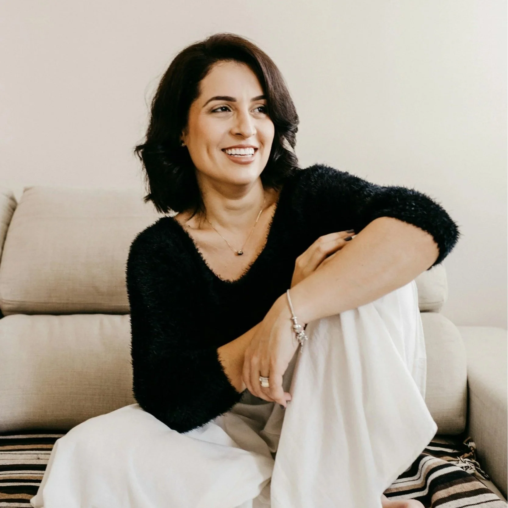 A woman with dark hair sitting on a beige sofa, smiling, wearing a black fuzzy top, silver jewelry, and striped socks.