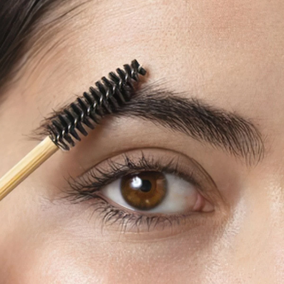 Close-up of a person with brown eye, applying mascara to their eyebrow with a spoolie brush.