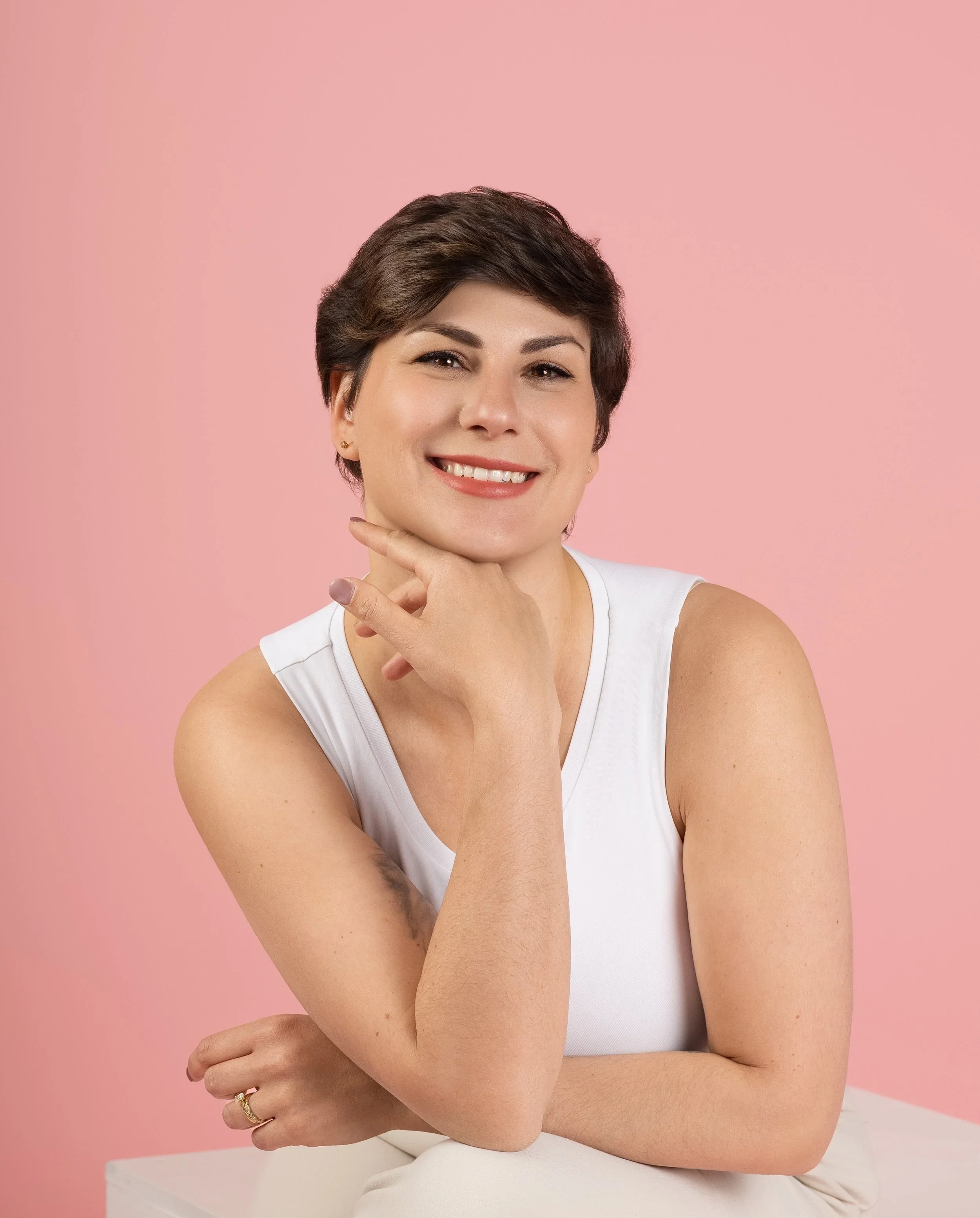 A woman with short brown hair smiling and resting her chin on her hand against a pink background.