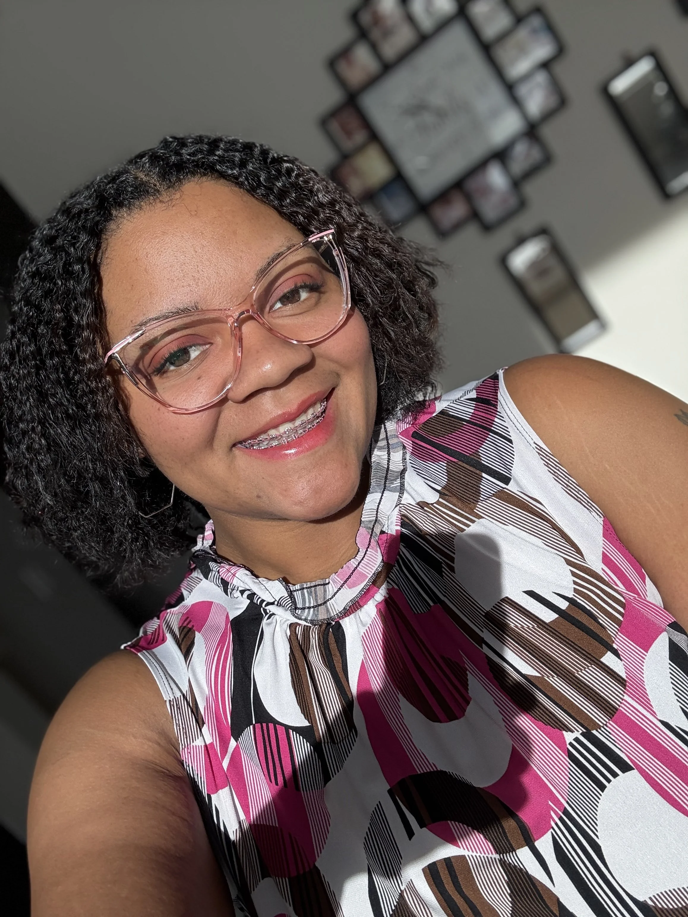 A woman with short, curly black hair wearing pink glasses and a sleeveless patterned top, smiling in front of a wall with a photo collage.