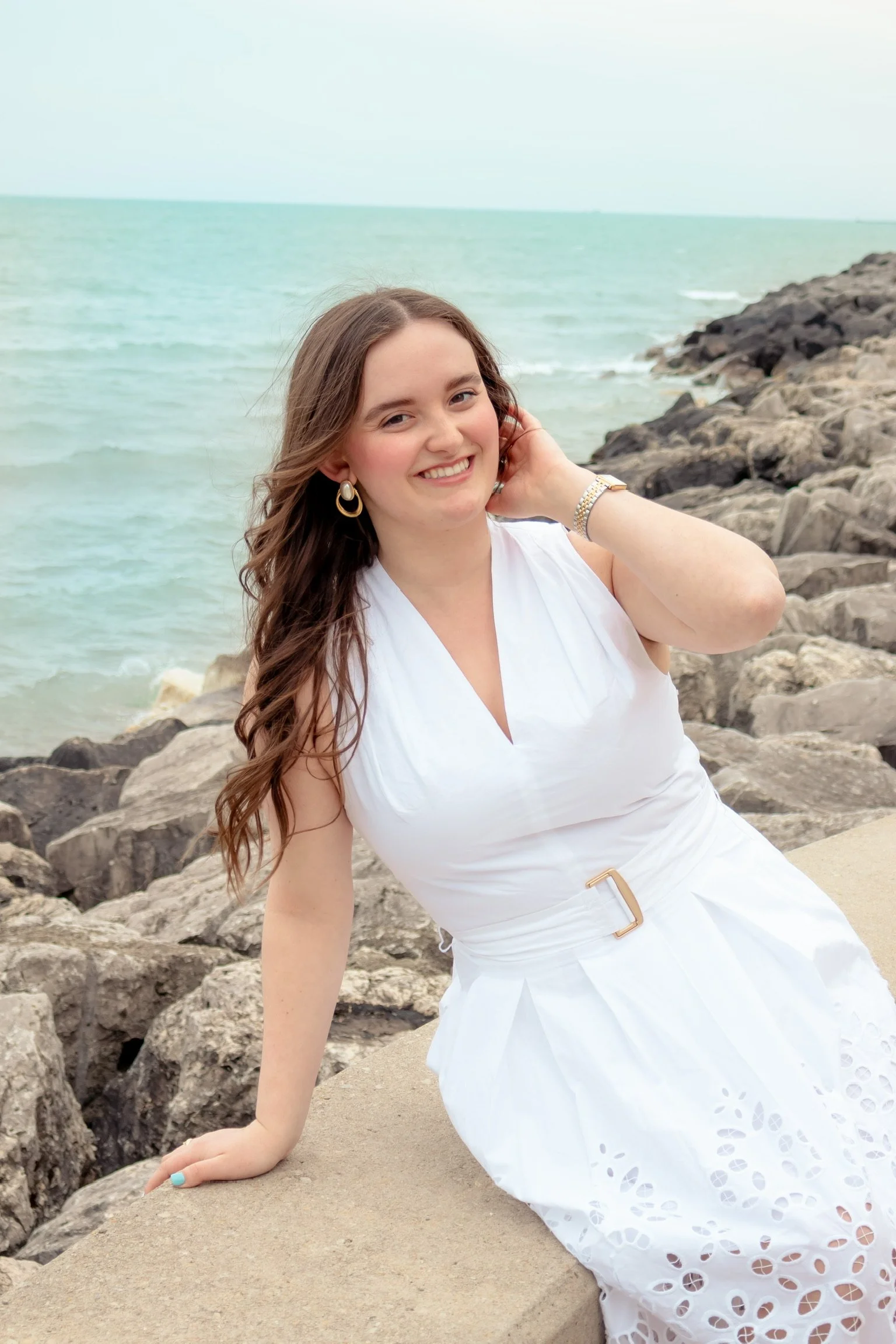 A woman with long brown hair, smiling in a white dress, sitting on a rock by the ocean with rocks in the background.