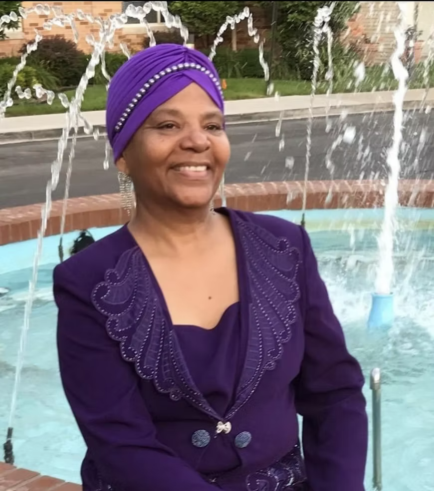 Woman with a purple headwrap and matching purple outfit smiling in front of a fountain with water spraying behind her.
