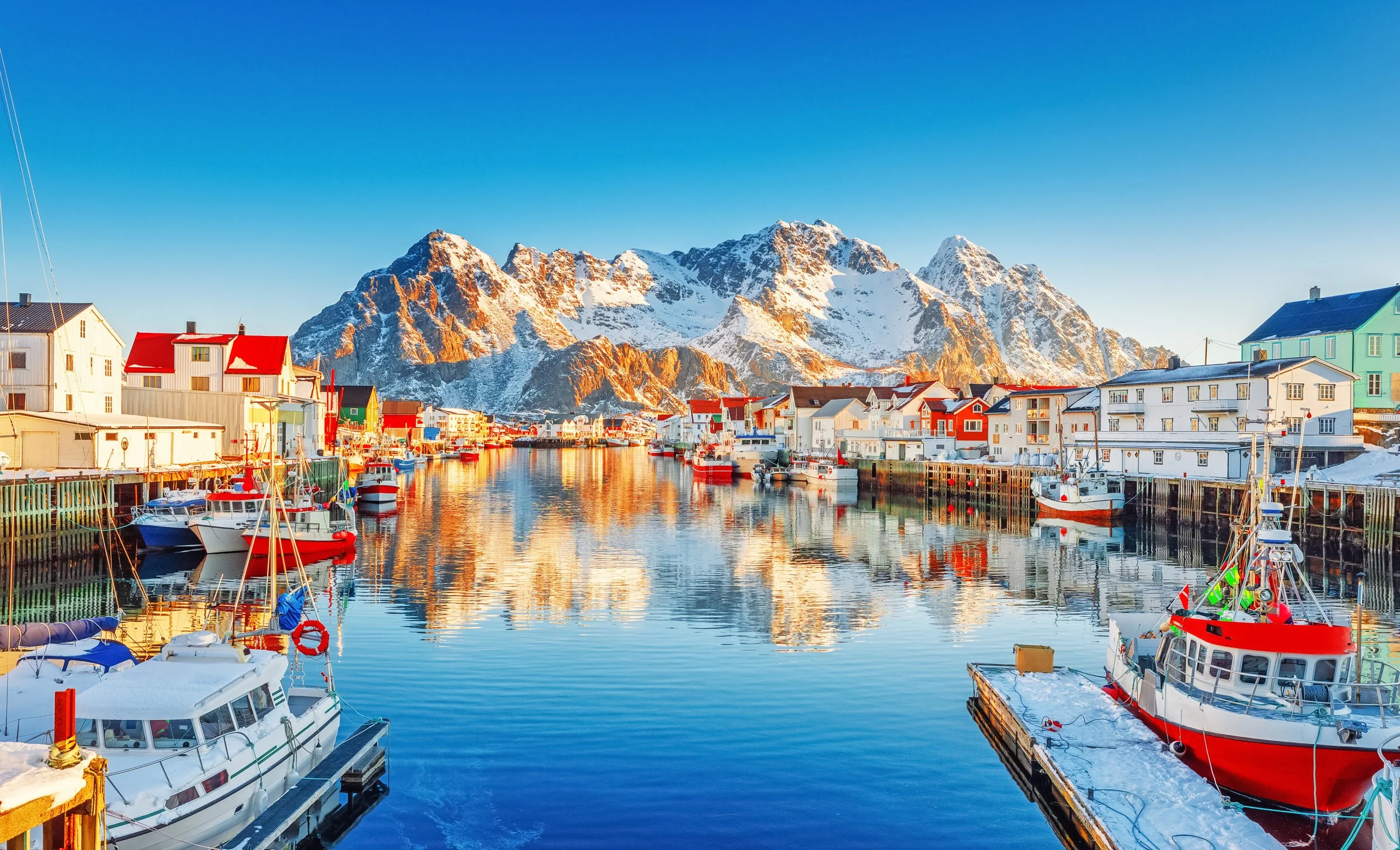A scenic coastal town with colorful houses and boats docked in a calm harbor, snow-capped mountains in the background, and clear blue skies.