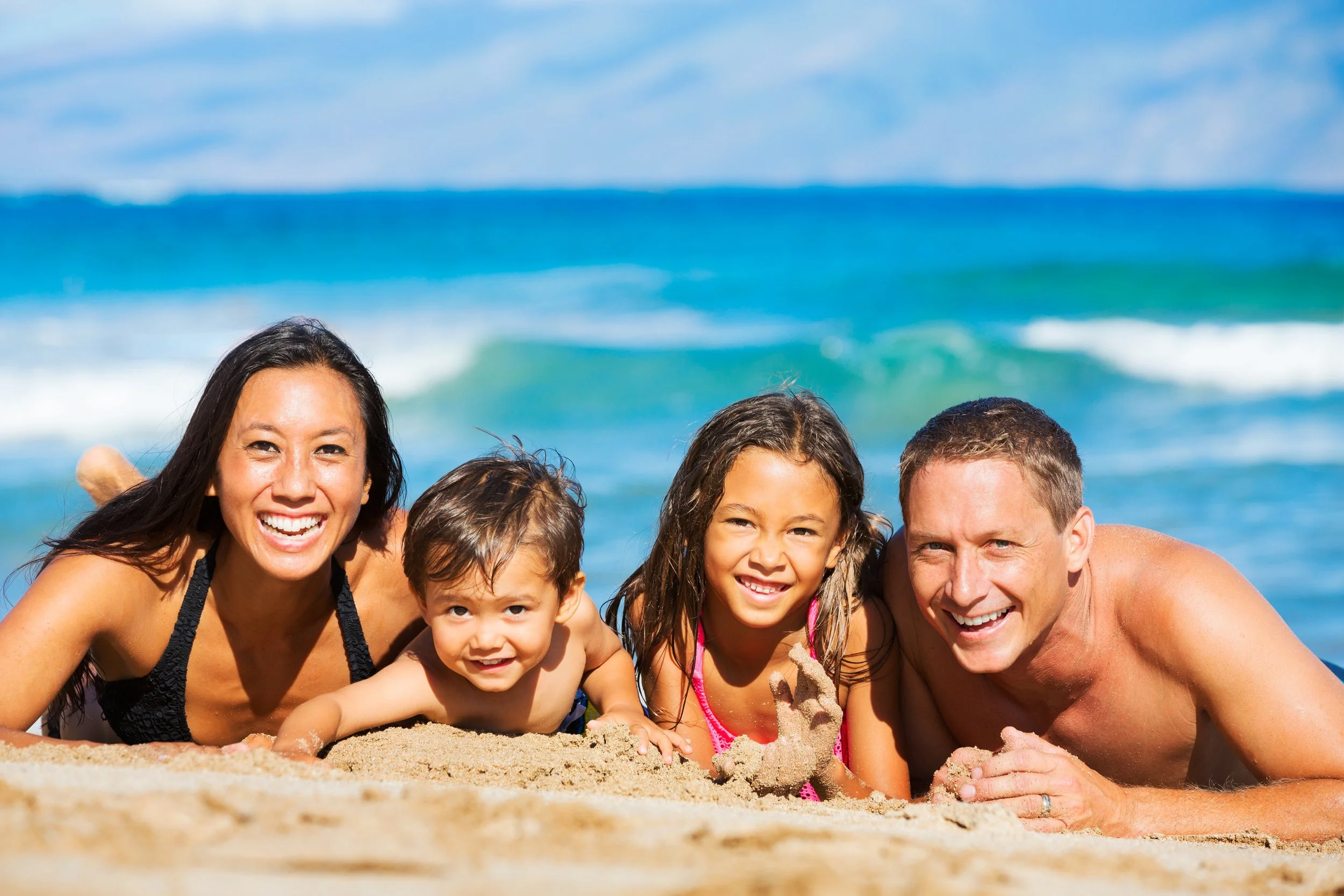 A happy family of four lying on the sand at the beach, smiling with the ocean in the background.