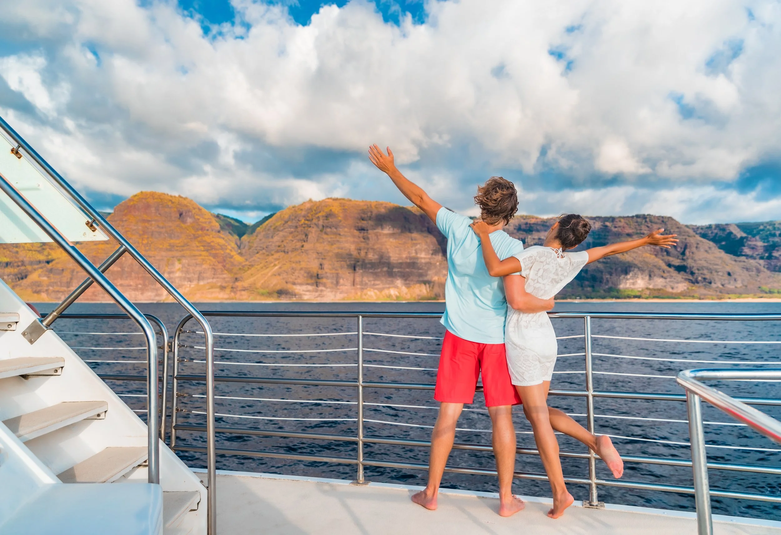 A couple on a boat enjoying a scenic view of mountains and water, with arms outstretched.