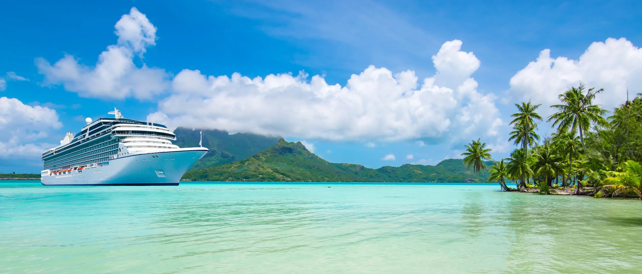 A large white cruise ship docked in a tropical bay with turquoise water, lush green mountains in the background, and palm trees on the right side under a partly cloudy sky.