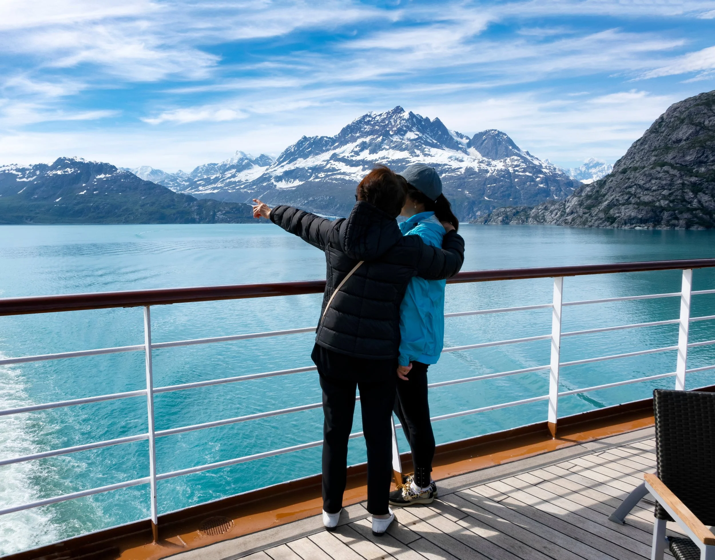 Two women stand on a ferry deck overlooking a turquoise lake with snow-capped mountains in the background. One woman points towards the mountains while the other woman leans into her.