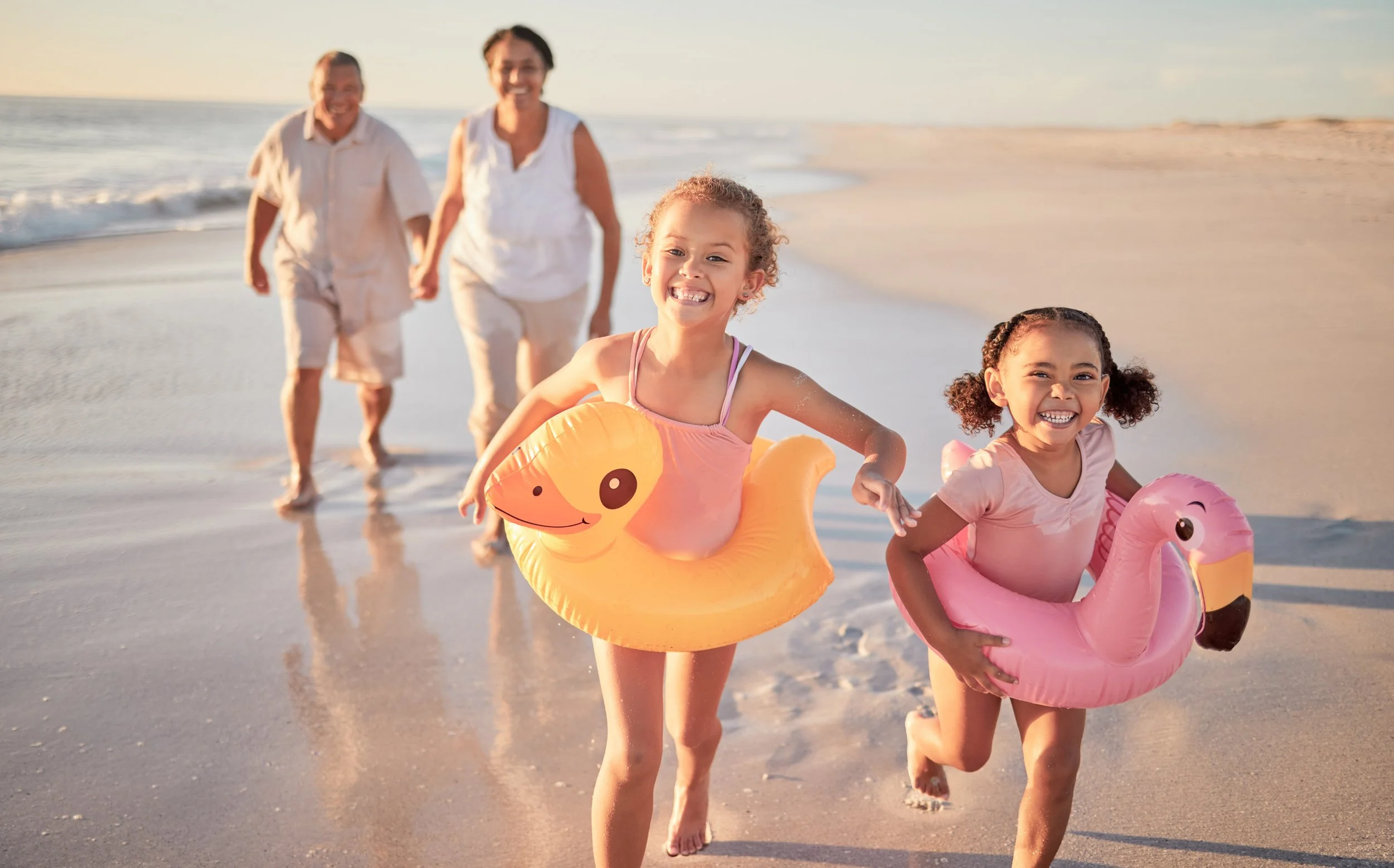 Two young girls running along the beach with inflatable pool floats, one shaped like a duck and the other like a flamingo, while two older generation adults walk behind them by the water at sunset.