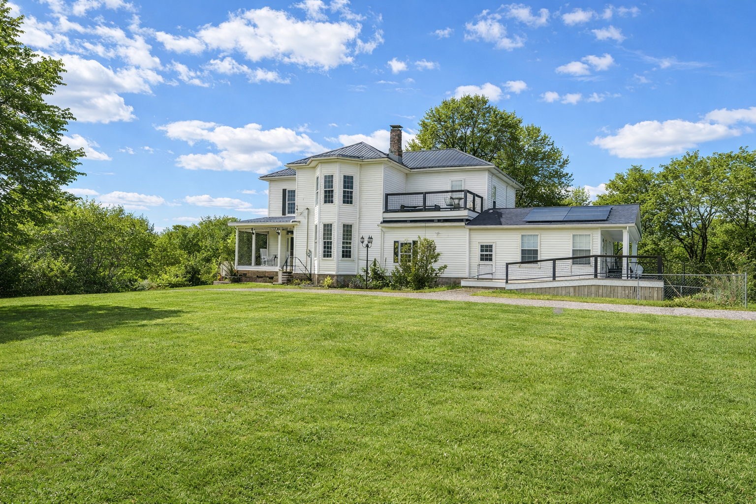A large white house with multiple levels and black railings sits on a lush green lawn under a blue sky with scattered clouds.