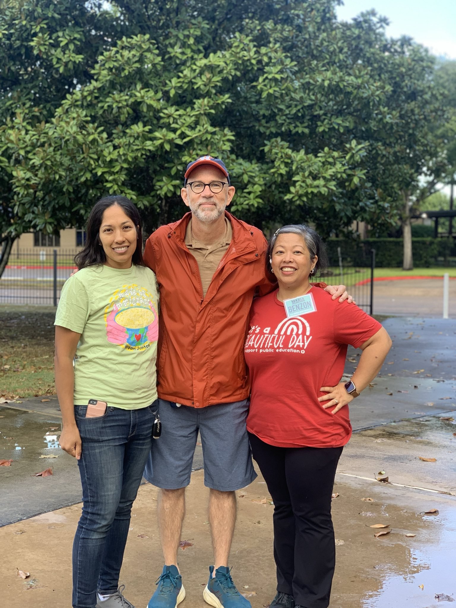 Three people standing outdoors in front of a tree, smiling, with a wet sidewalk and patches of leaves around them.