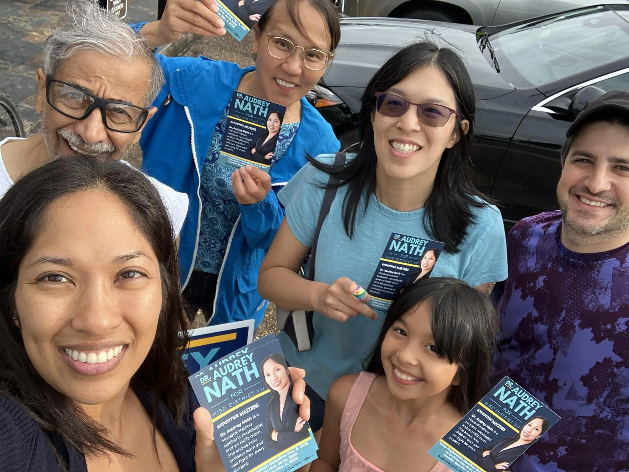 Group of people gathered outdoors, smiling and holding campaign flyers and a brochure for Dr. Audrey Nath, who is running for office in District C. There are men, women, and a young girl in the group, standing in front of a parked car.