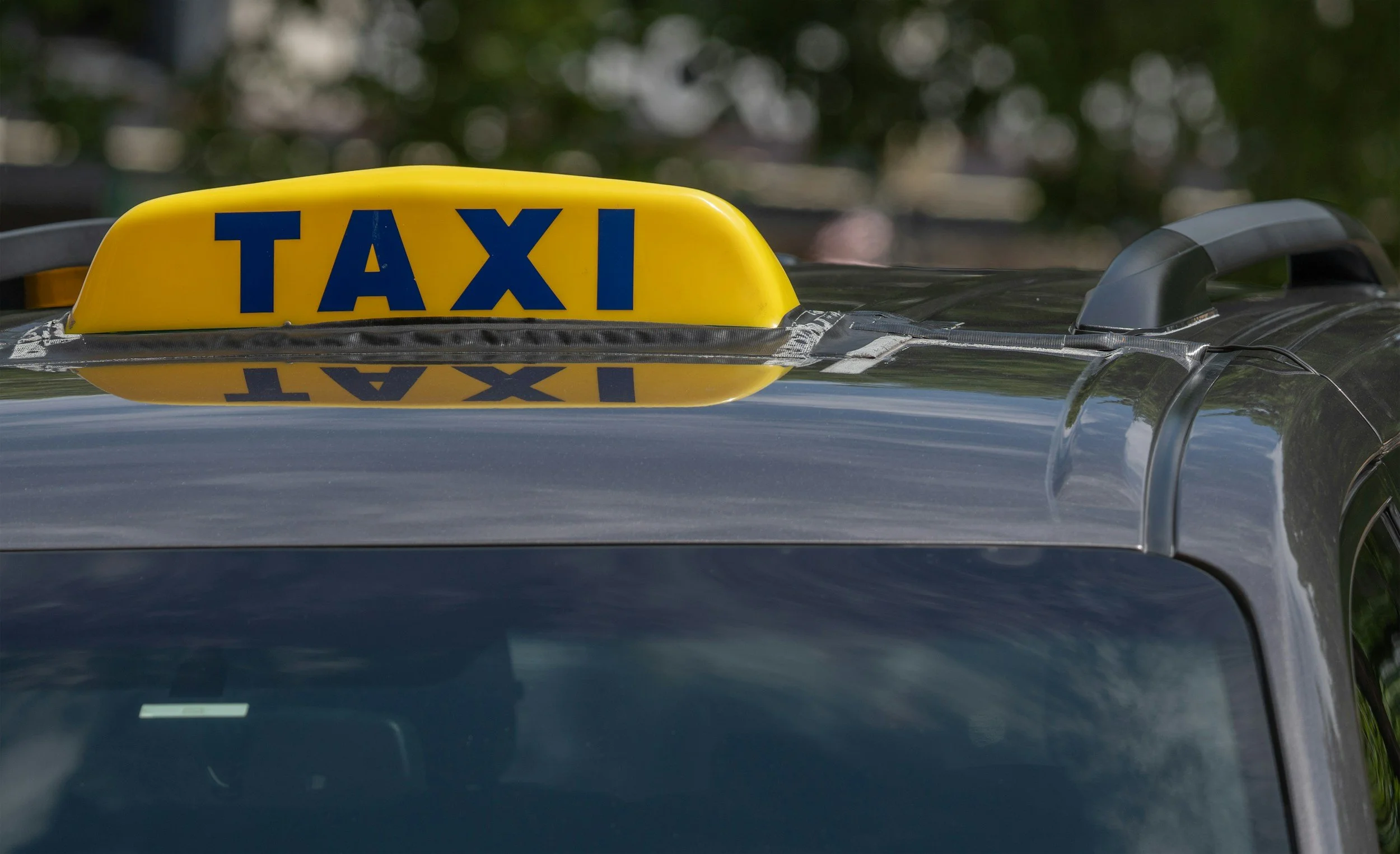 Close-up of a yellow taxi sign on top of a black taxi car, with the word 'TAXI' written in blue letters, reflecting on the car's roof.