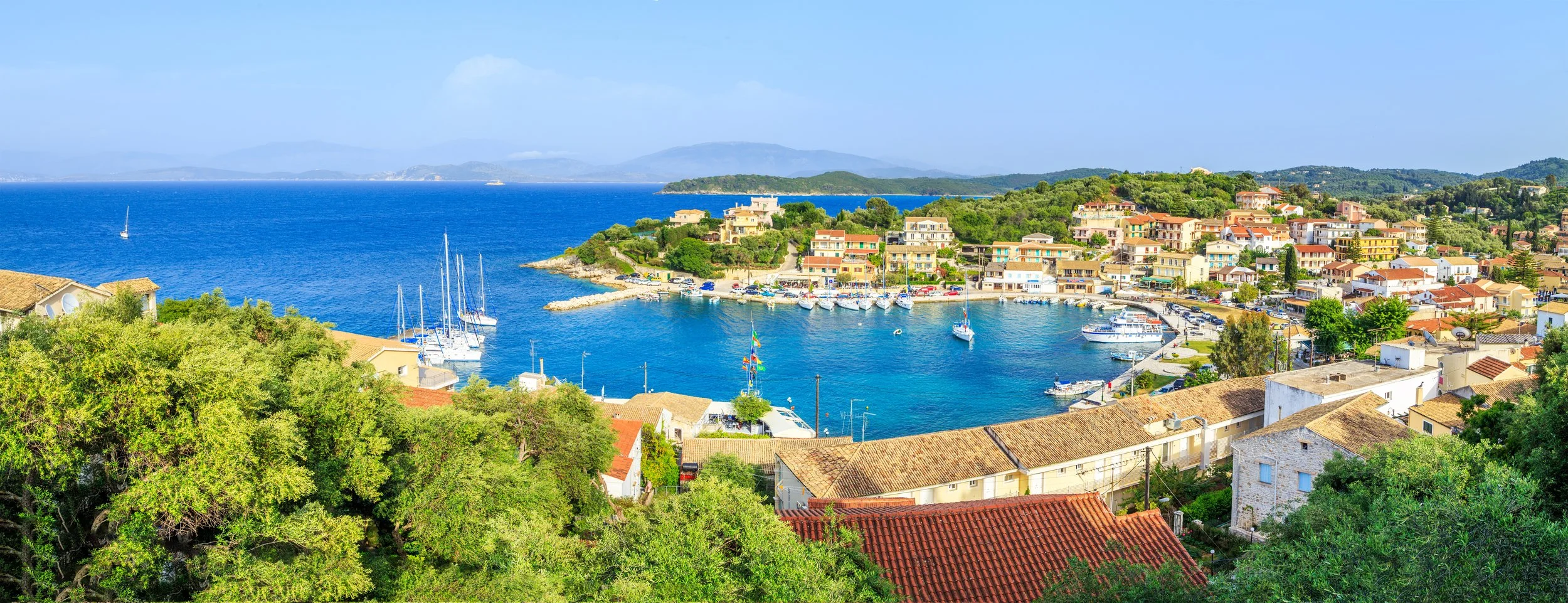 Kassiopi harbour in north east Corfu with boats, colourful village buildings and Ionian Sea views.