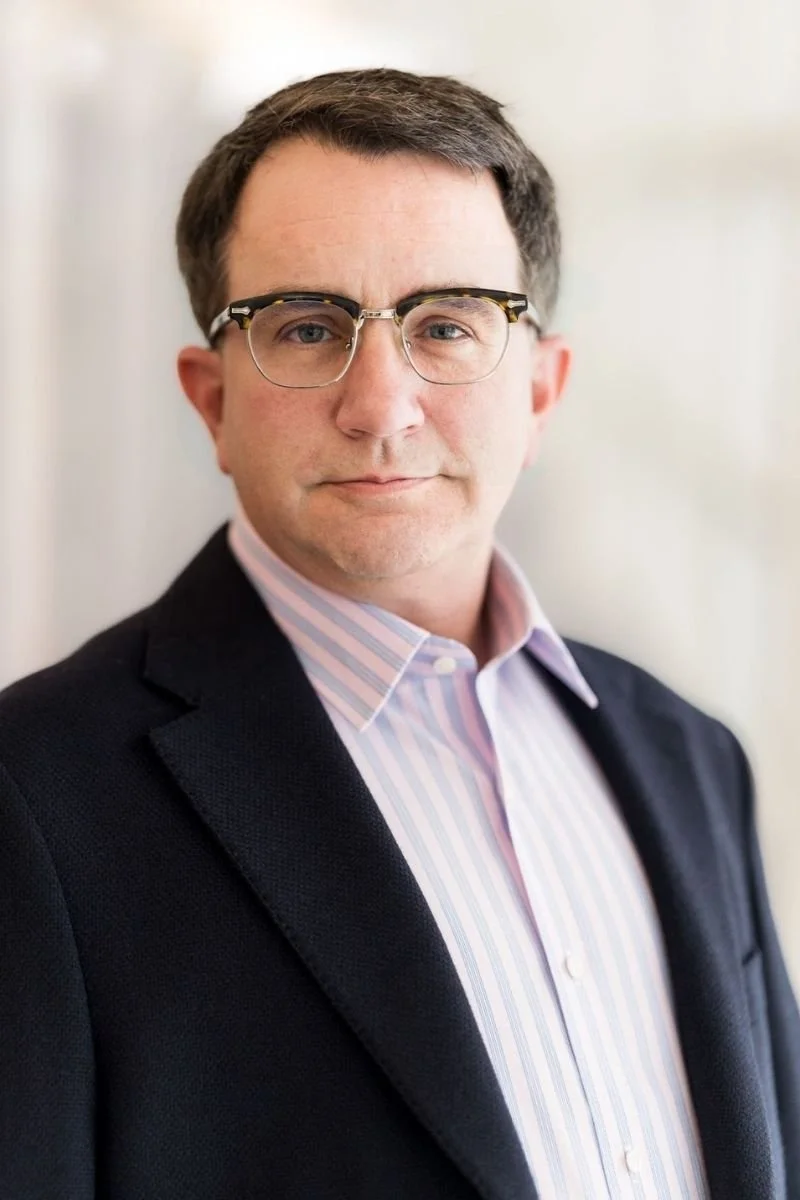 A man with dark hair and glasses in a suit jacket and a light striped pink and white shirt standing indoors.