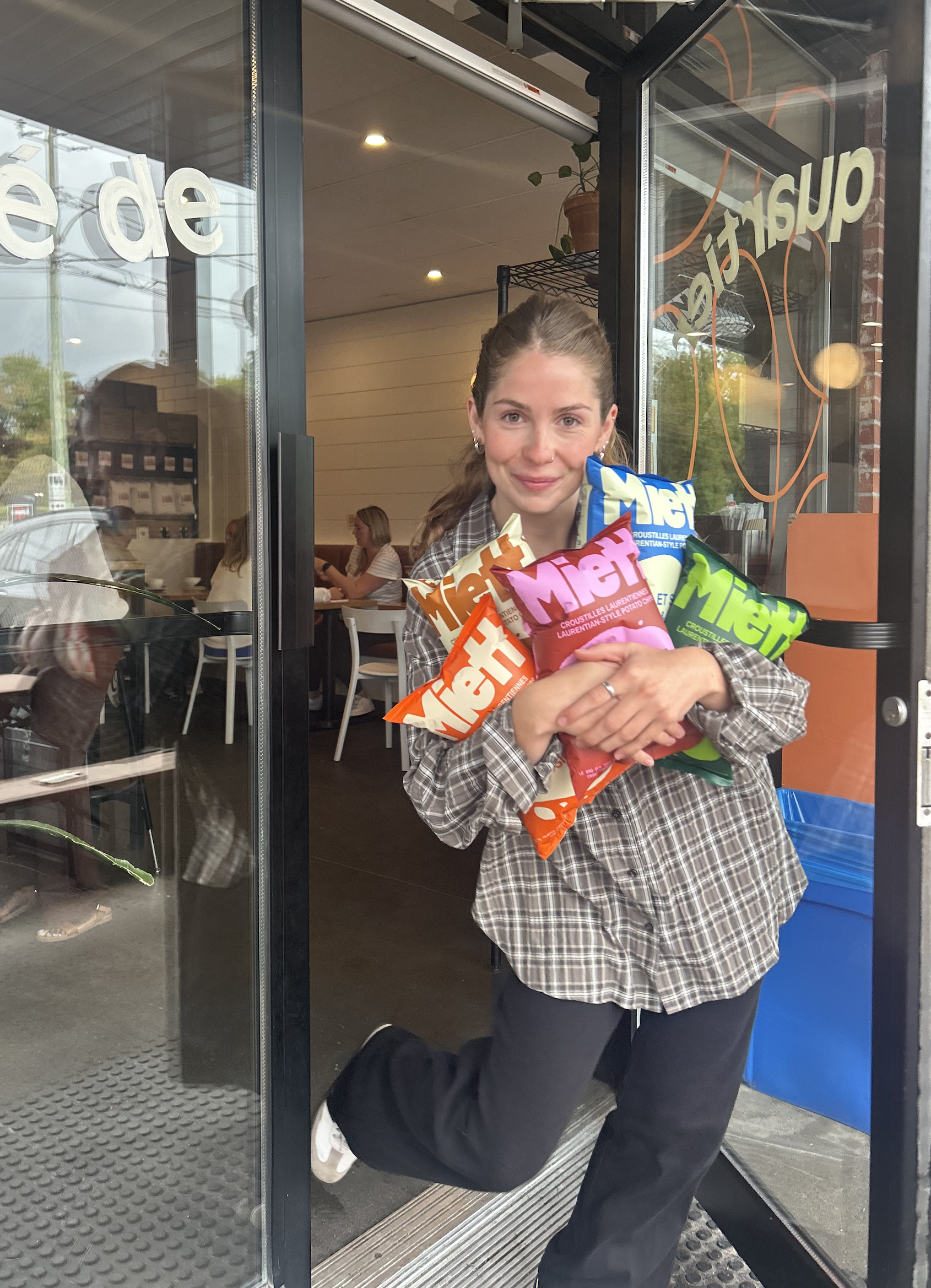 Une femme sortant d'un restaurant ou d'un café, tenant plusieurs paquets de snacks de la marque Mert, face à la porte d'entrée. Elle a les cheveux longs attachés, porte une chemise à carreaux et sourit.