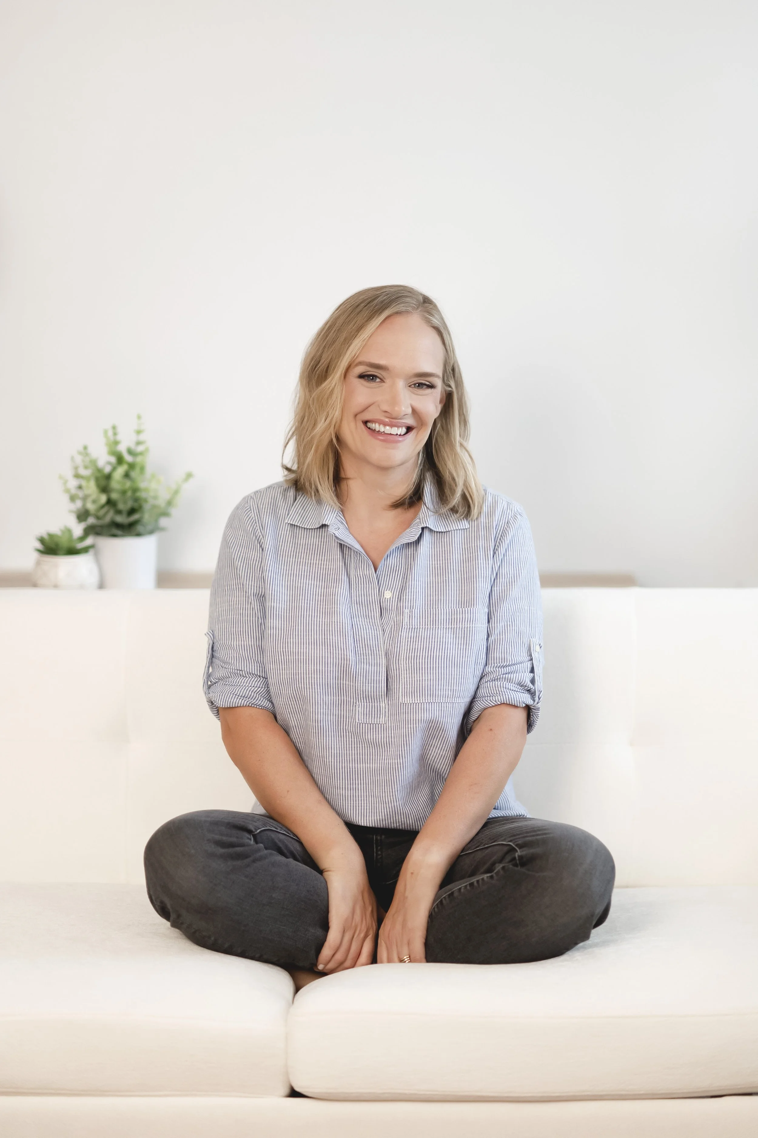 A woman sitting cross-legged on a white sofa, smiling, wearing a light blue striped shirt and dark jeans, with a potted plant in the background.