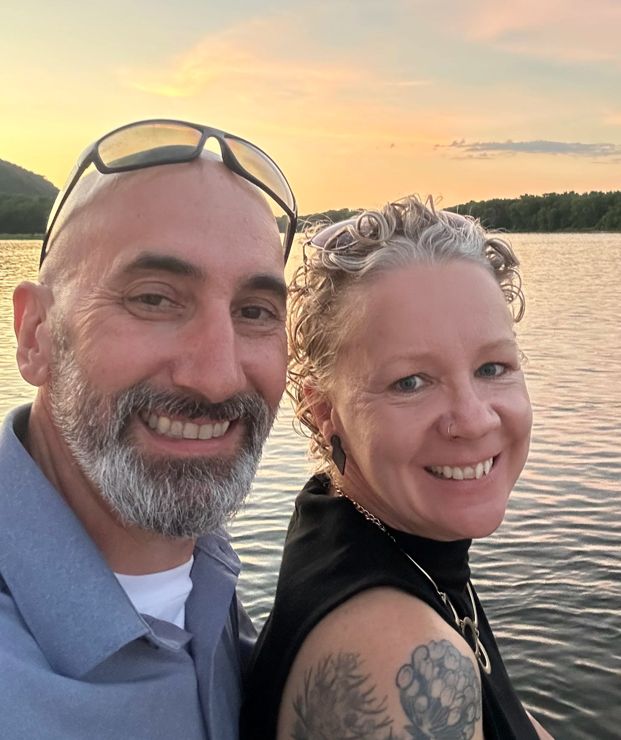 A smiling couple taking a selfie by a river during sunset.