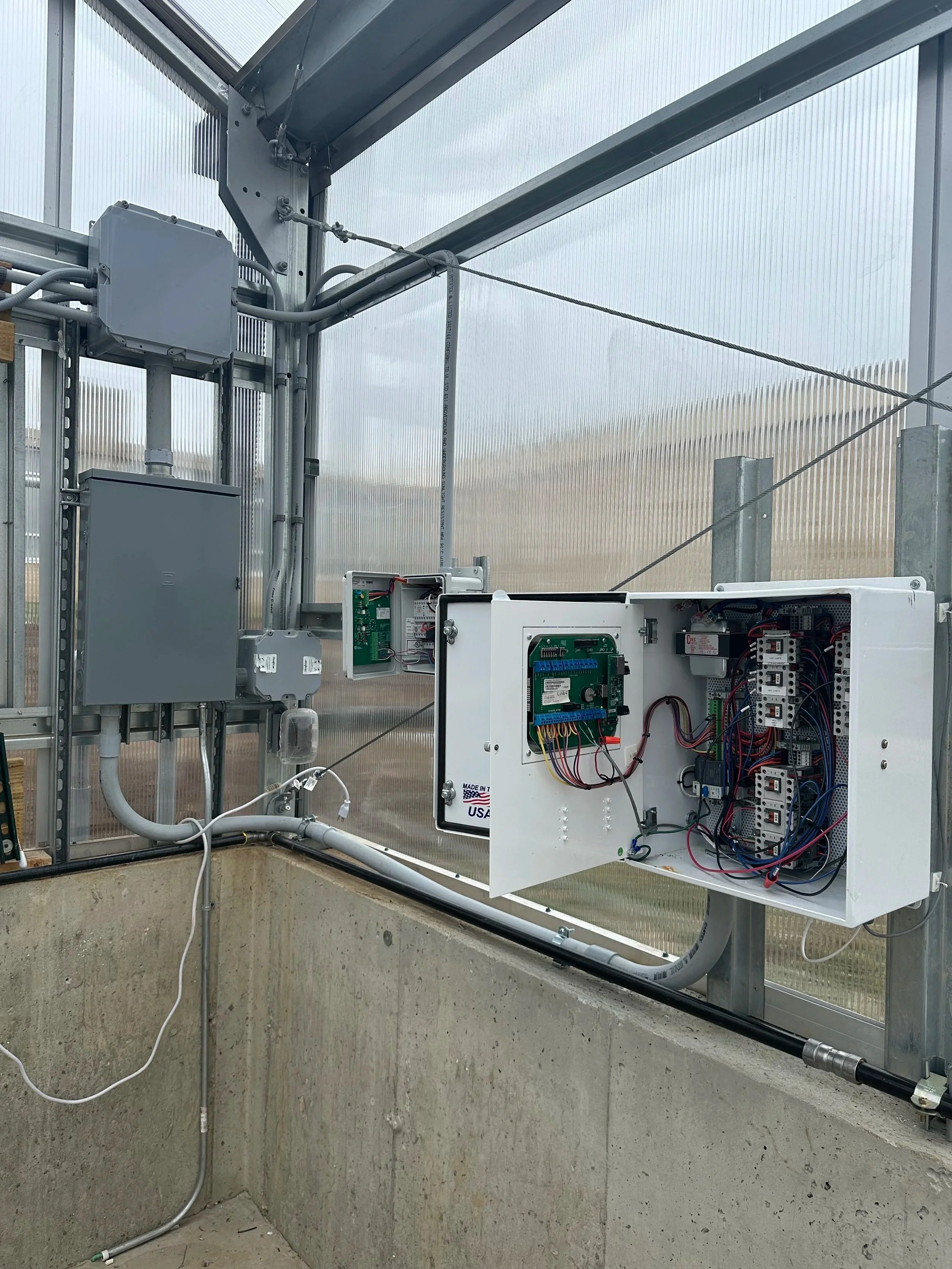 Small electrical cabinet with contactors and control wiring inside mounted to wall on interior of a greenhouse.