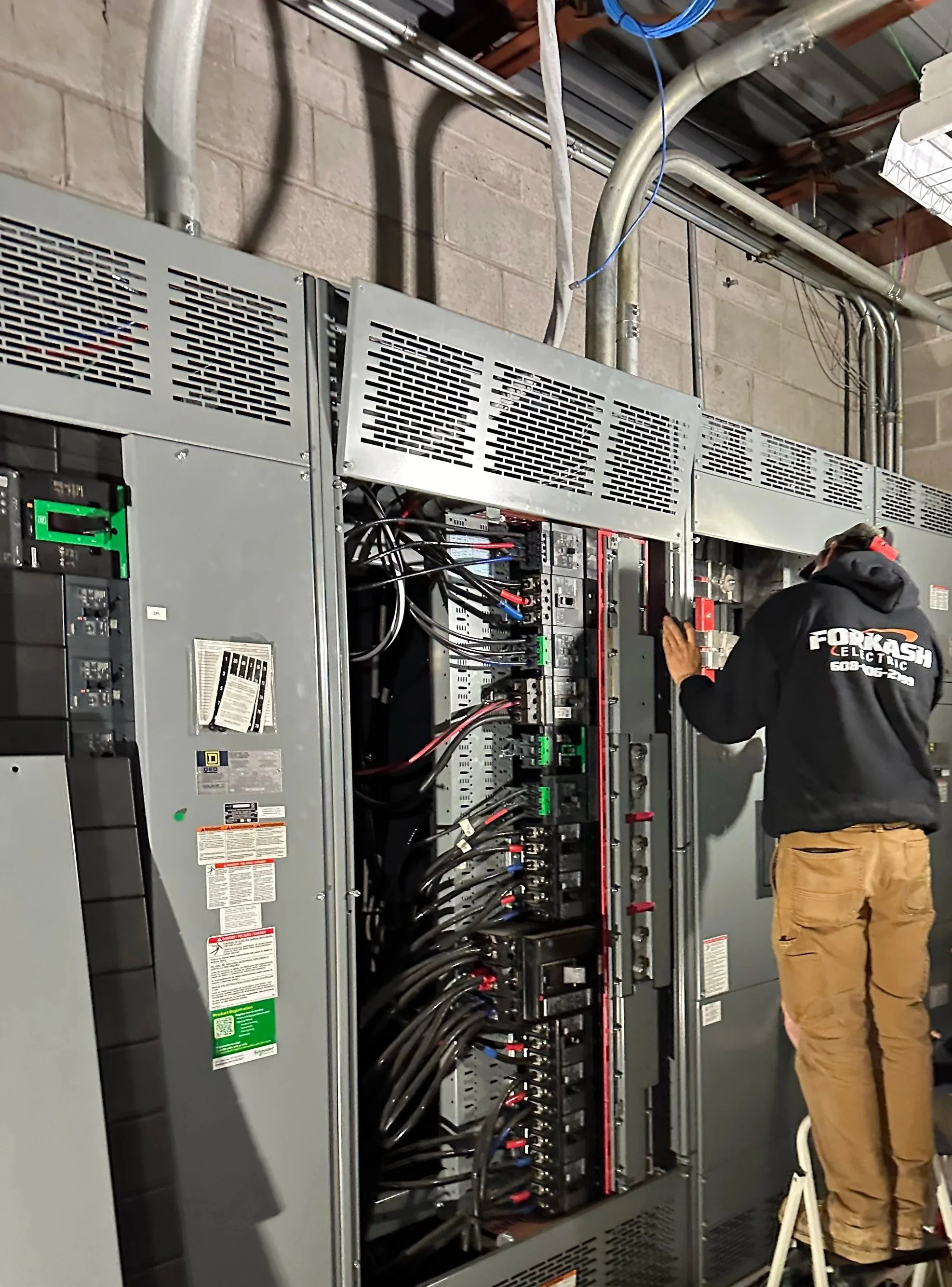 Electrician working on power distribution equipment consisting of several grey metal cabinets, large circuit breakers, and multiple wires.