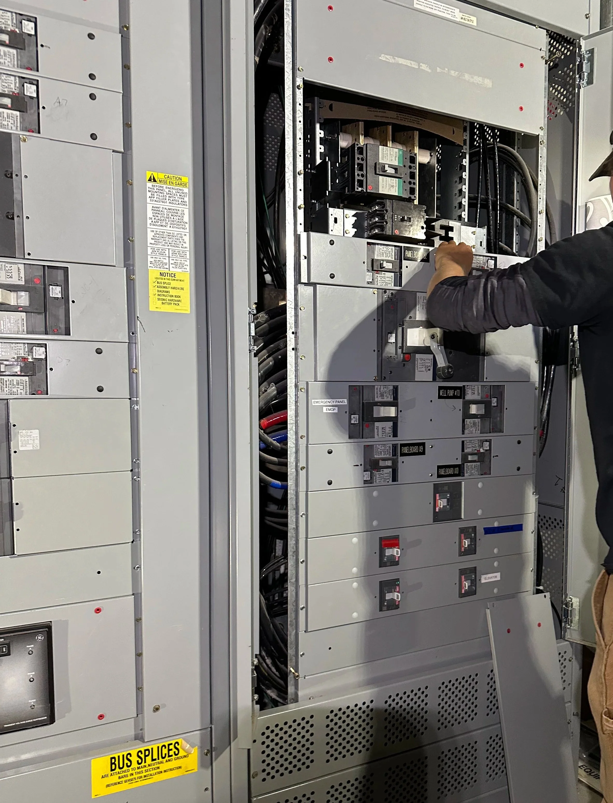 Electrician working on large gray metal cabinet filled with many circuit breakers and disconnects.