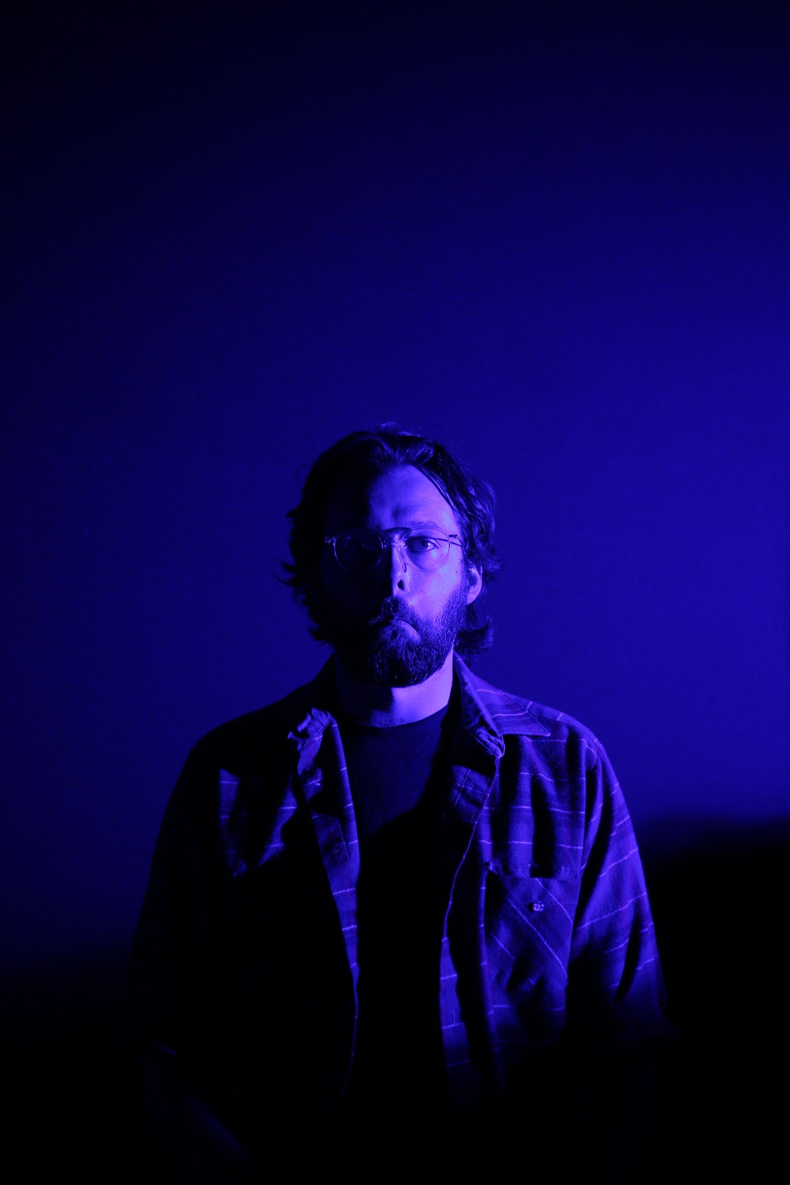 A man with glasses and a beard, wearing a checkered shirt, is illuminated by blue and purple light against a dark background.