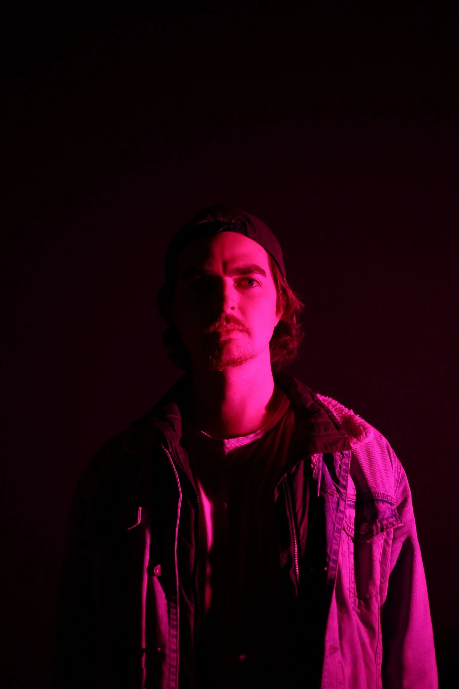 Portrait of a young man with long hair, wearing a backwards cap and a jacket, illuminated by pink low-key lighting against a dark background.