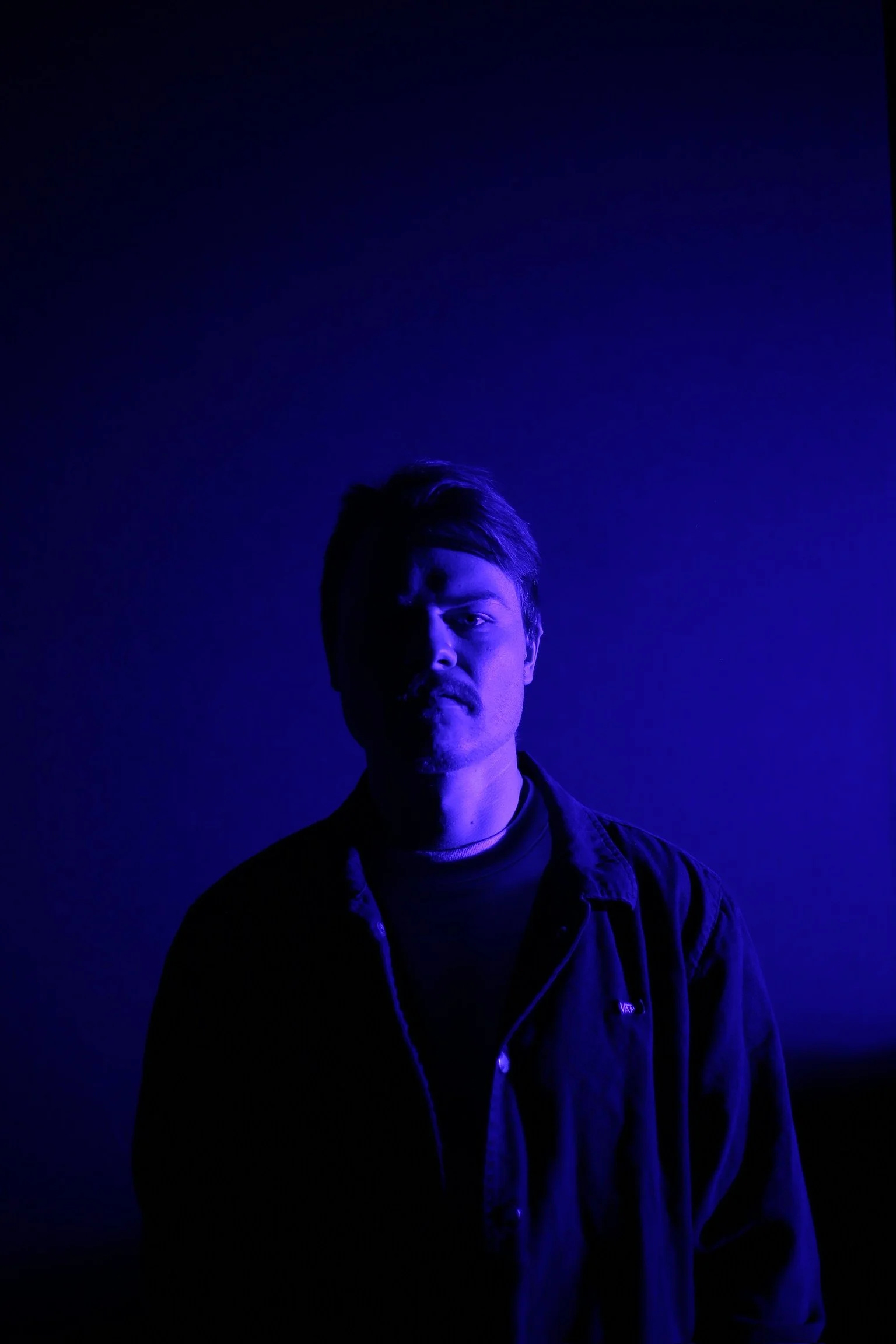 A portrait of a man in blue and purple lighting, facing forward with a serious expression, wearing a dark jacket over a shirt.
