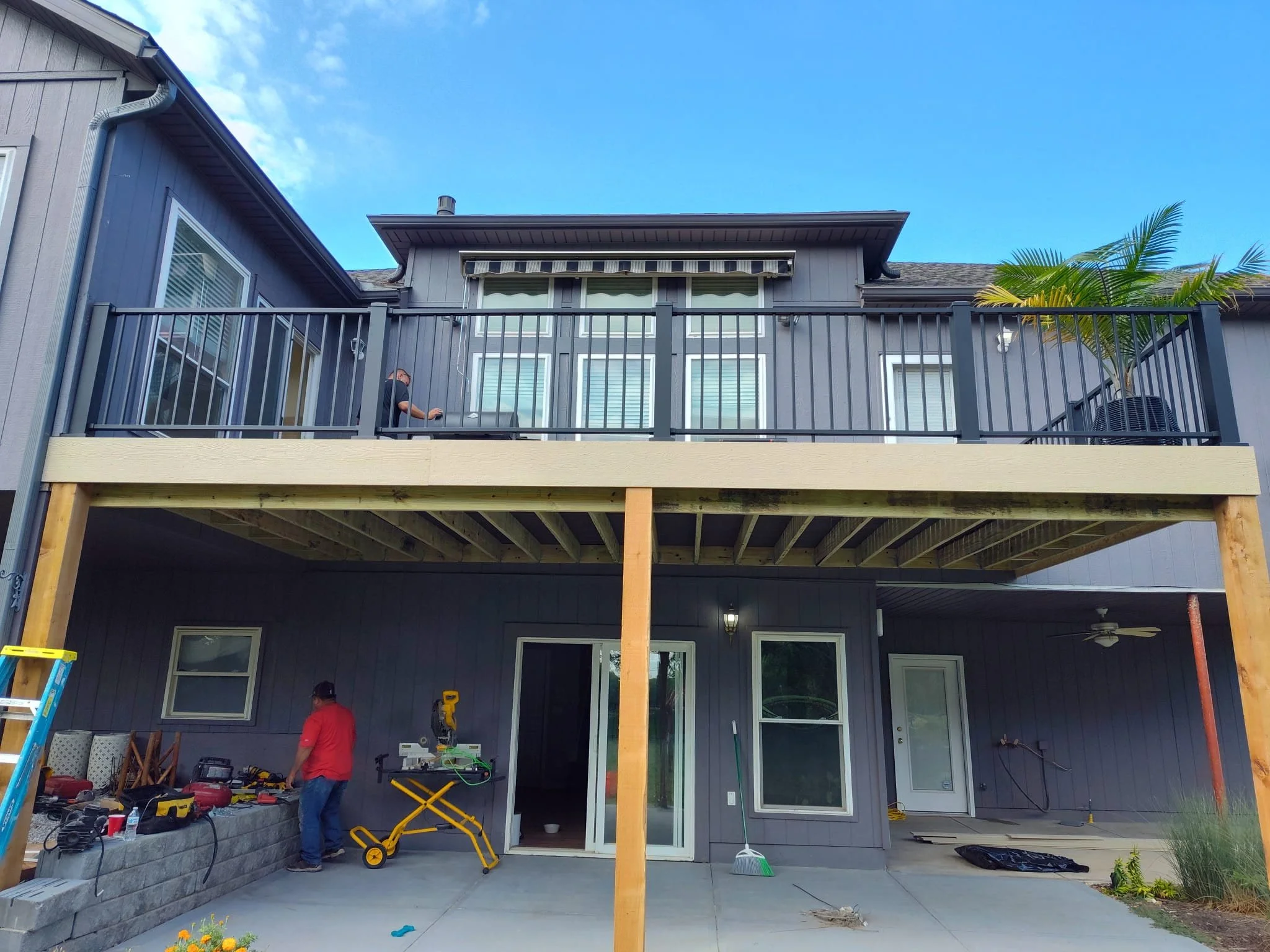 View of a two-story house with a newly constructed balcony. The upper balcony has black metal railing and a person working on it, with a palm tree visible in the corner. The lower level shows a patio area with tools and two people working, one wearin