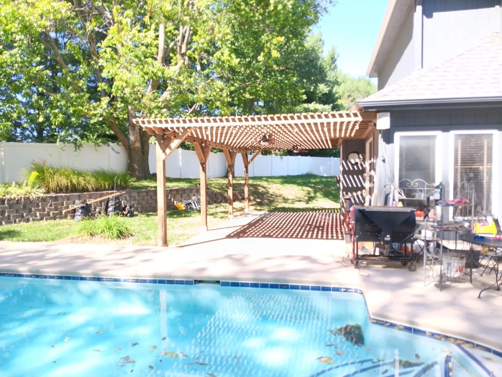 Backyard with swimming pool, wooden shade structure, lush trees, and patio furniture.