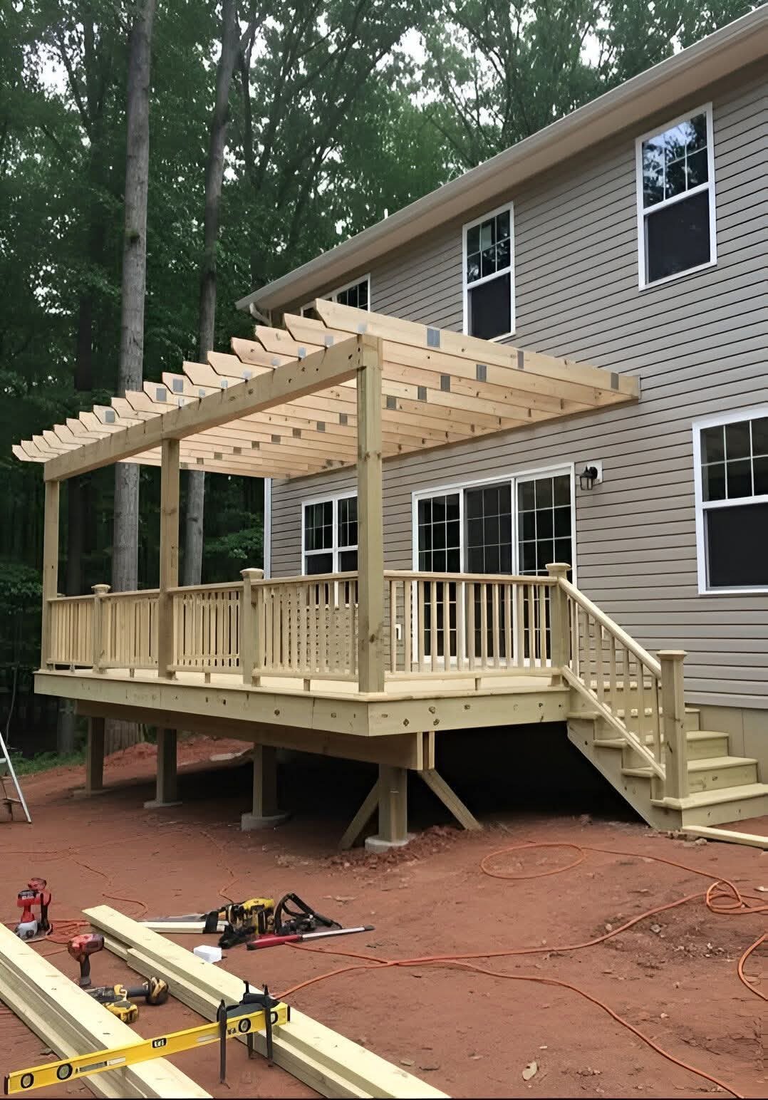A wooden deck under construction attached to a beige house with multiple windows, surrounded by trees, with construction tools and materials in the foreground.