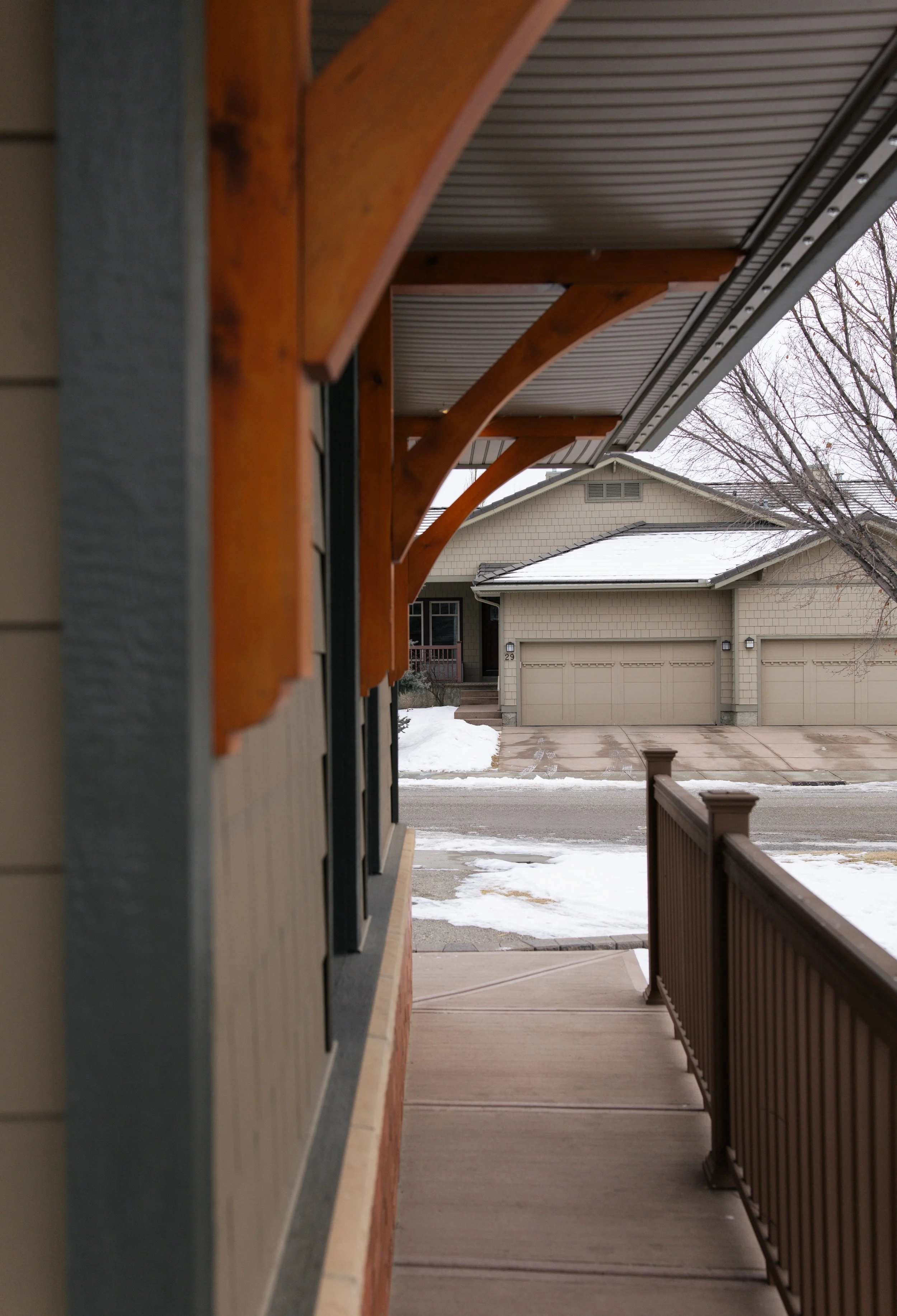 View from a porch showing a neighboring house with a two-car garage, snow on the ground, leafless trees, and a wet sidewalk.