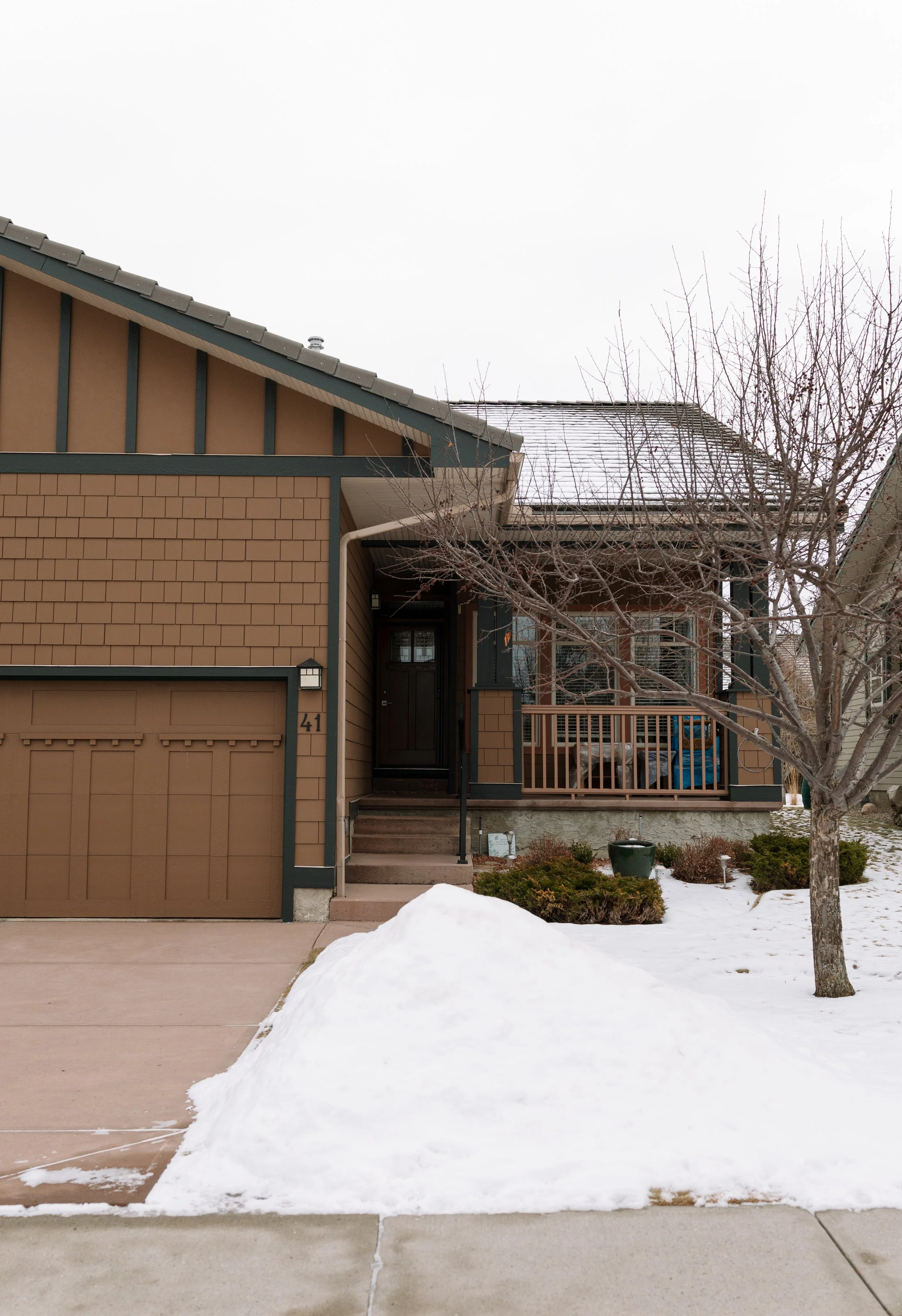 A two-story brown house with a garage, front steps, and a small porch area, with a snow-covered yard and leafless tree in front on a cloudy winter day.