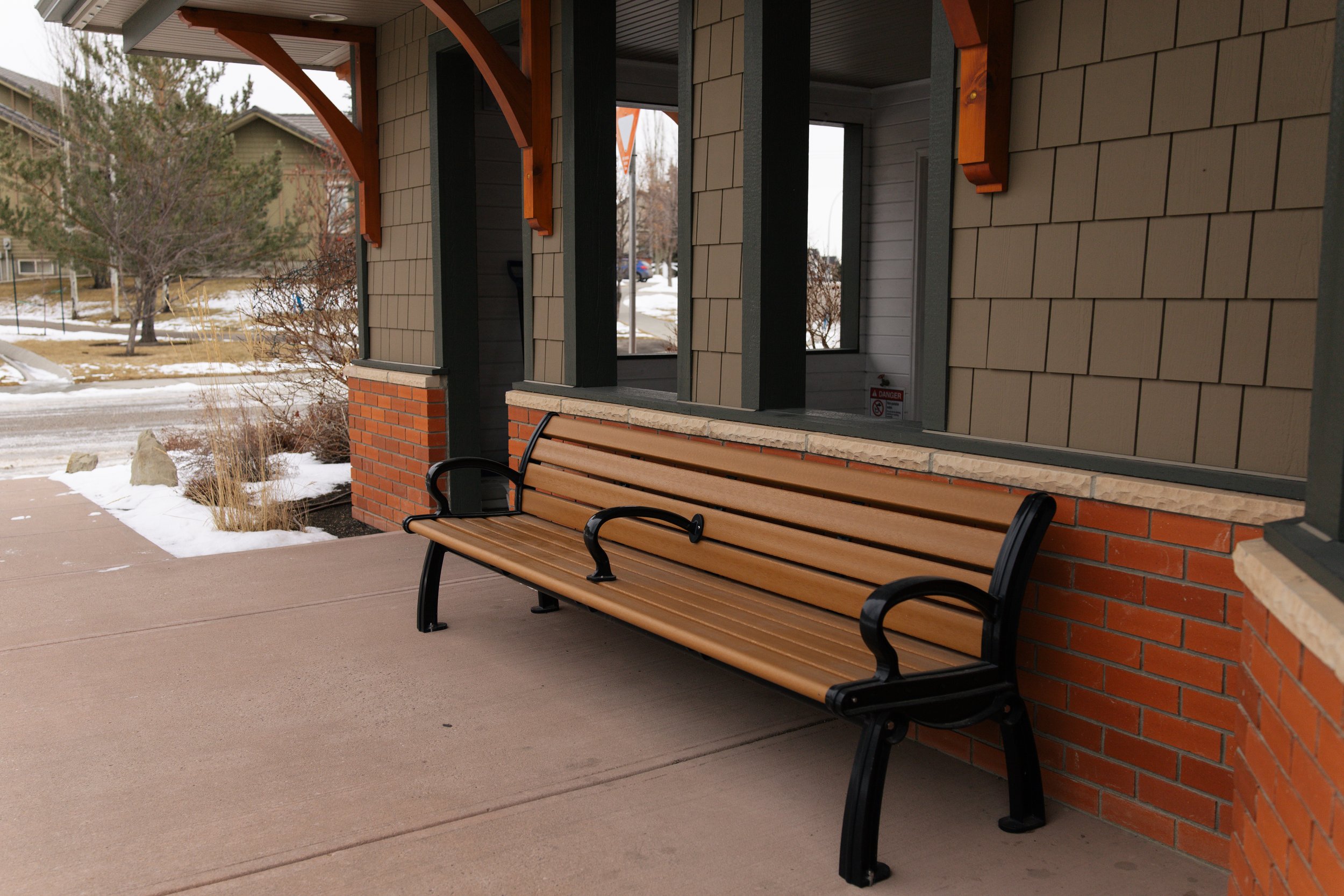 Empty wooden bench on the sidewalk outside a building with brick and gray siding during winter.