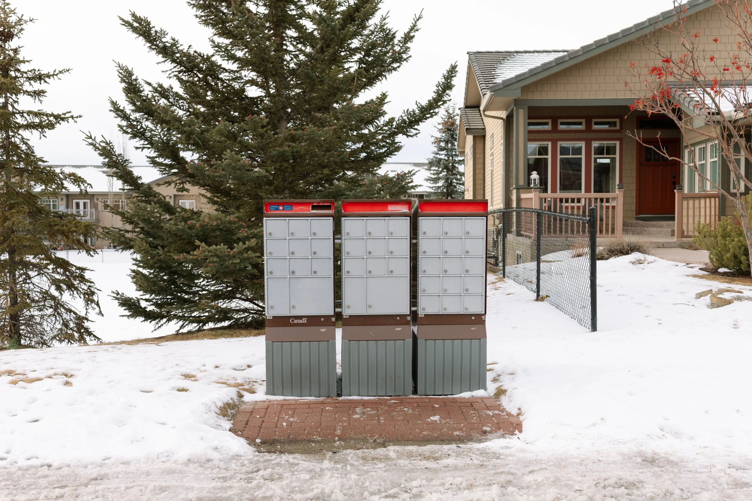 Three mailbox units on a snow-covered sidewalk in front of a house with a porch and trees in the background.